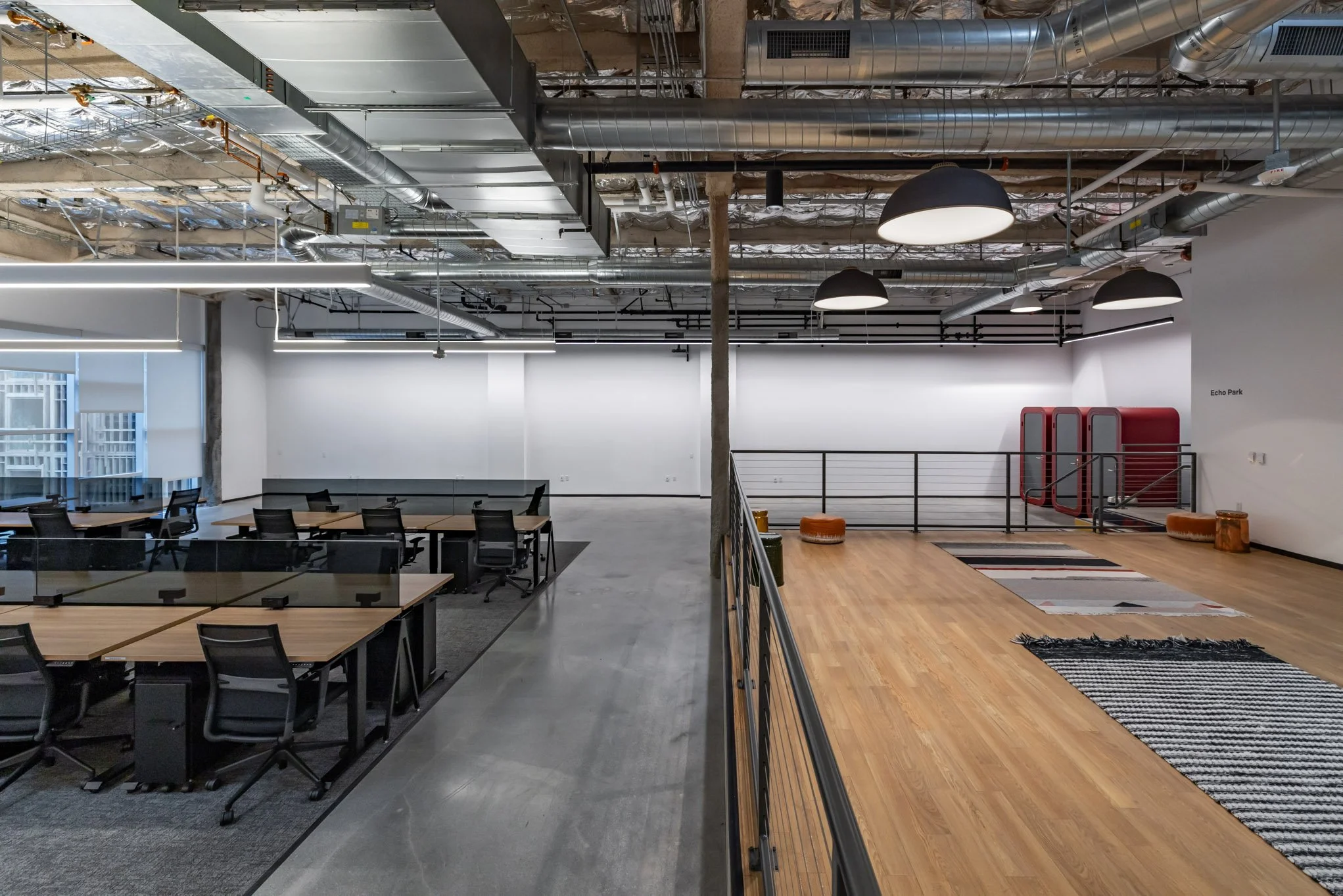 Partially divided modern office space with a conference room on the left, featuring desks and chairs, and an open lounge area on the right with rugs, poufs, and fitness equipment, under exposed ceiling pipes and large ceiling lights.