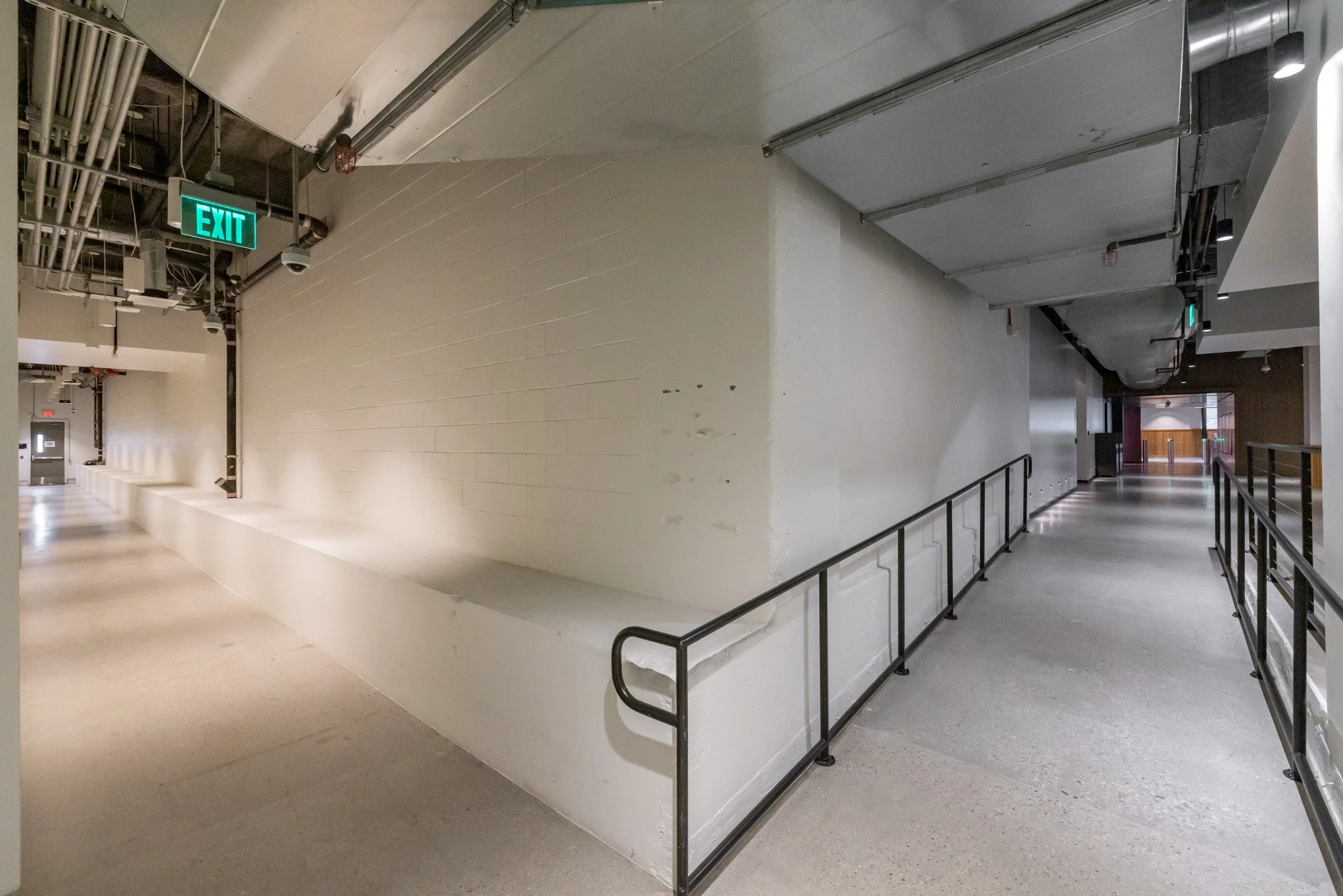 Empty indoor corridor with an exit sign, black safety railings, and visible ventilation pipes on ceiling.