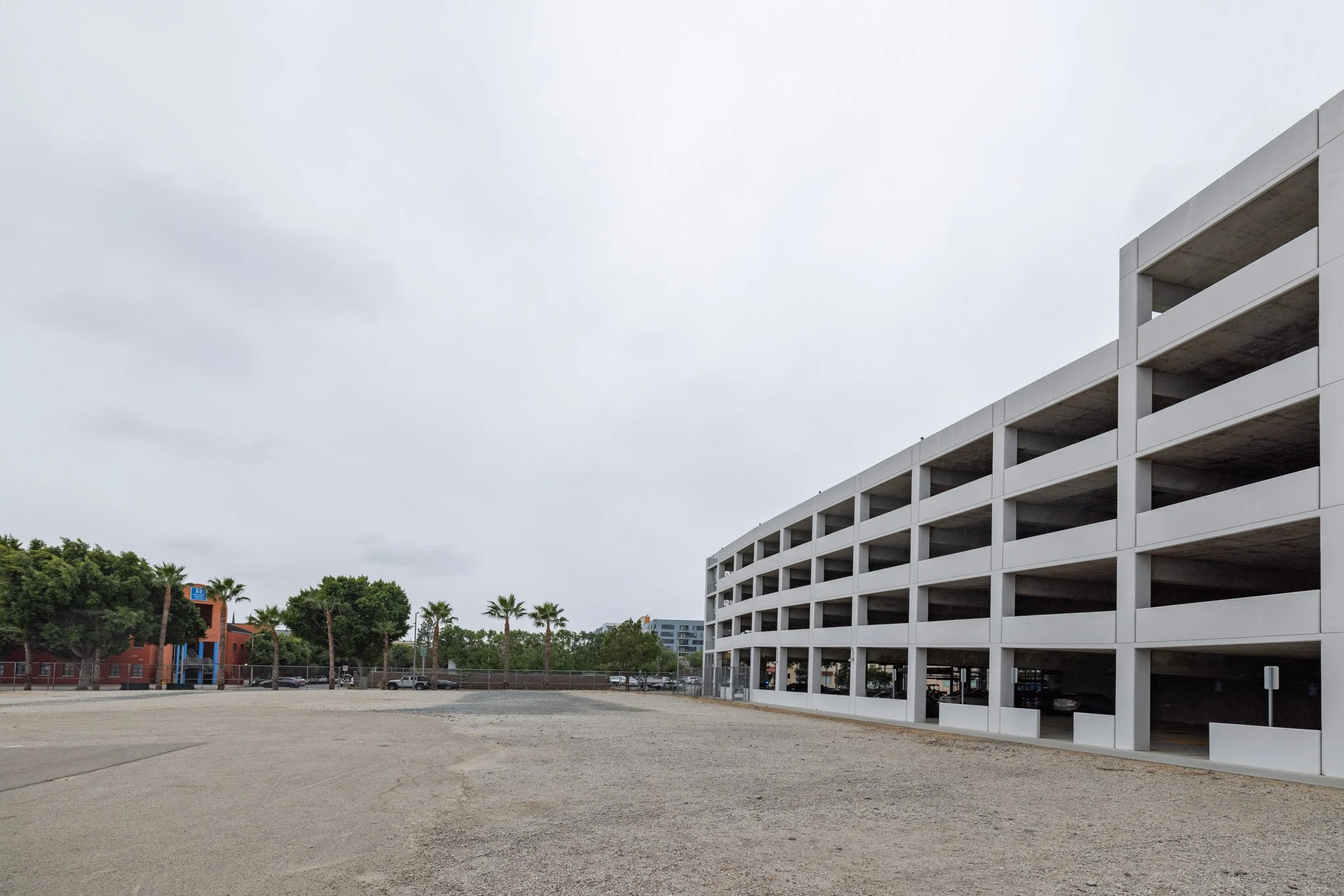 Empty outdoor parking garage with multiple levels, adjacent to a gravel lot, under an overcast sky.