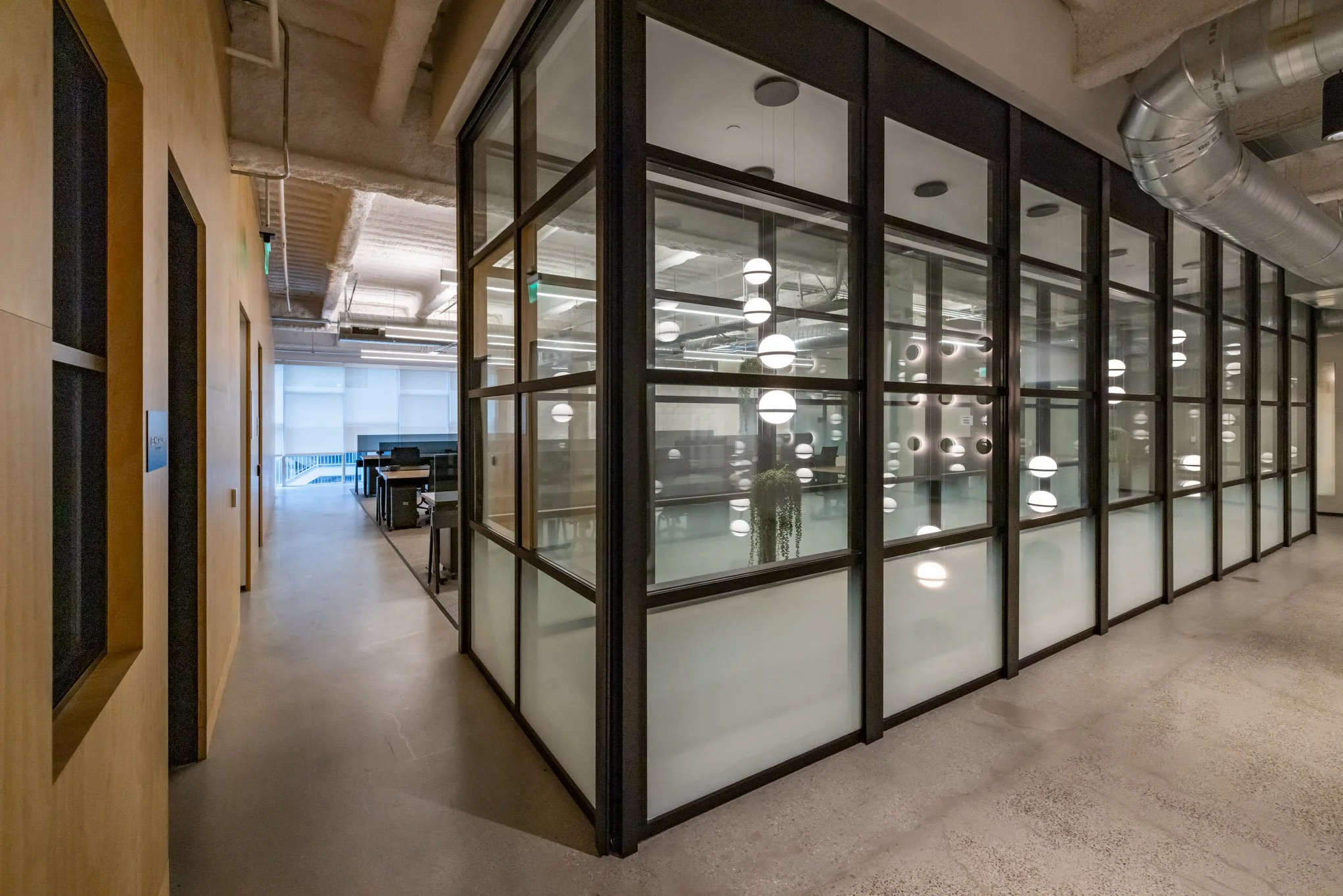 Modern office space with a glass-walled meeting room and a hallway leading to workstations, exposed ceiling pipes, and contemporary lighting.