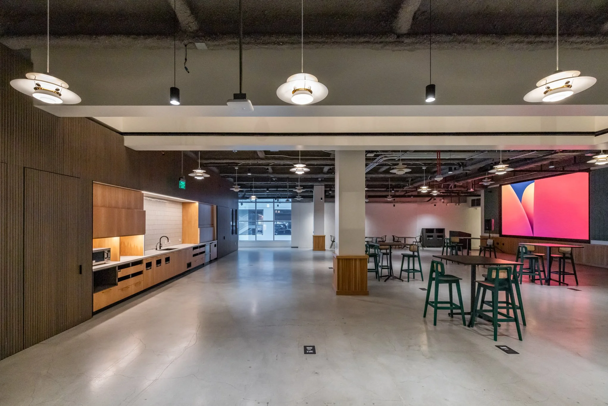 Modern interior of a spacious lounge or break room with tables, stools, a kitchenette, and a large digital screen on the wall.