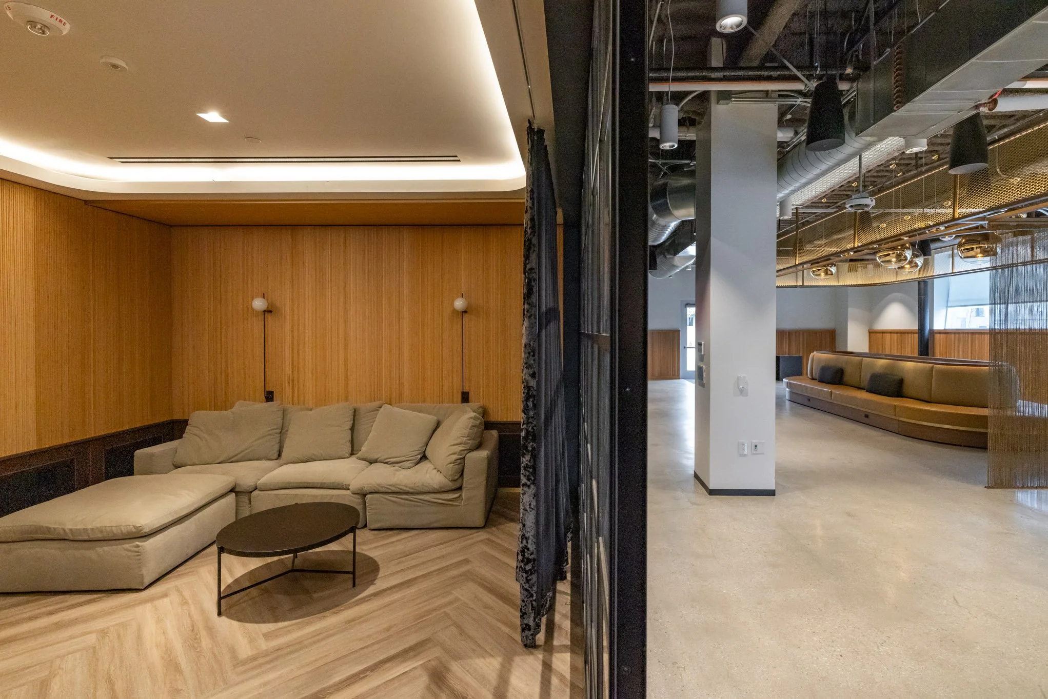 Two-tone interior with a beige sectional sofa, black coffee table, wooden wall panels, and off-white ceiling on the left; modern lounge with curved seating, wood panels, and exposed ceiling ducts on the right.