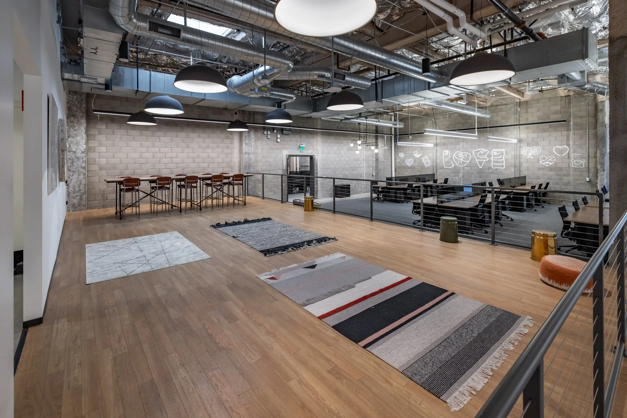 An open office space with a wooden floor, rugs, a long high table with chairs, lounge seating, modern lighting, exposed ductwork, and neon ice cream and food graphics on a wall.