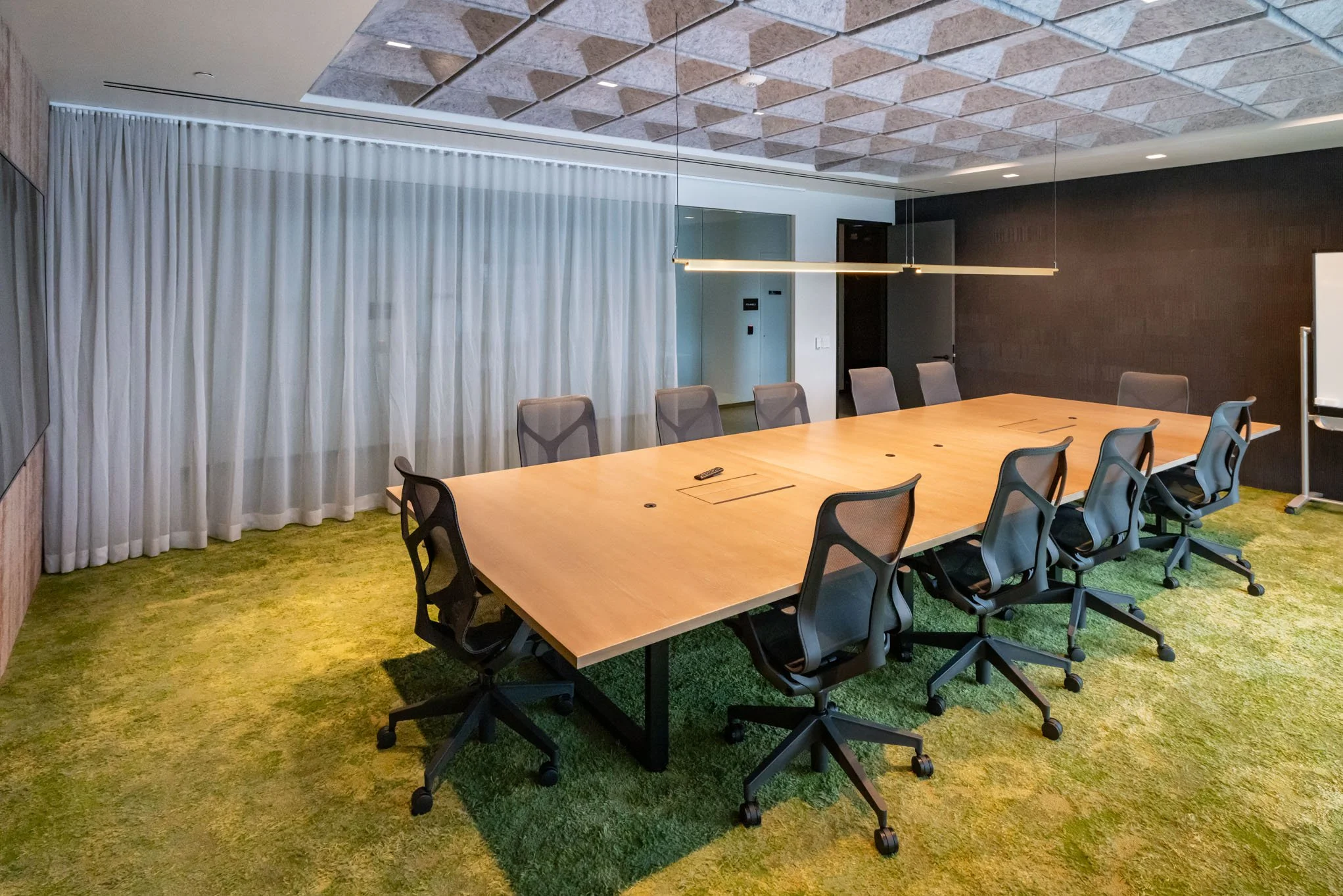 Empty modern conference room with a large wooden table, ten gray office chairs, green carpet, white curtain, and minimal wall decorations.