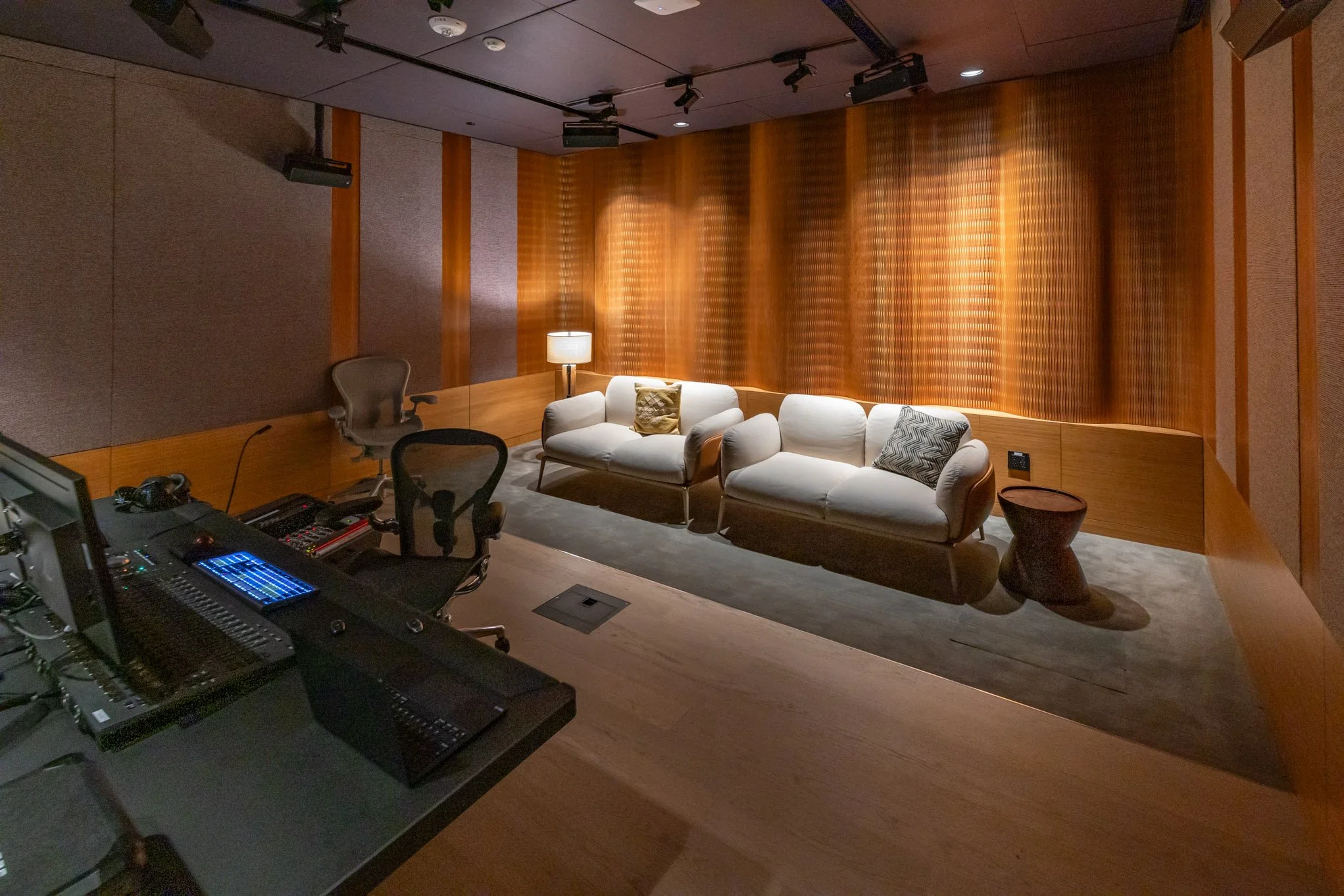A recording studio with two white sofas, a side table, a floor lamp, and audio equipment on a desk, with wood-paneled walls and spotlights overhead.