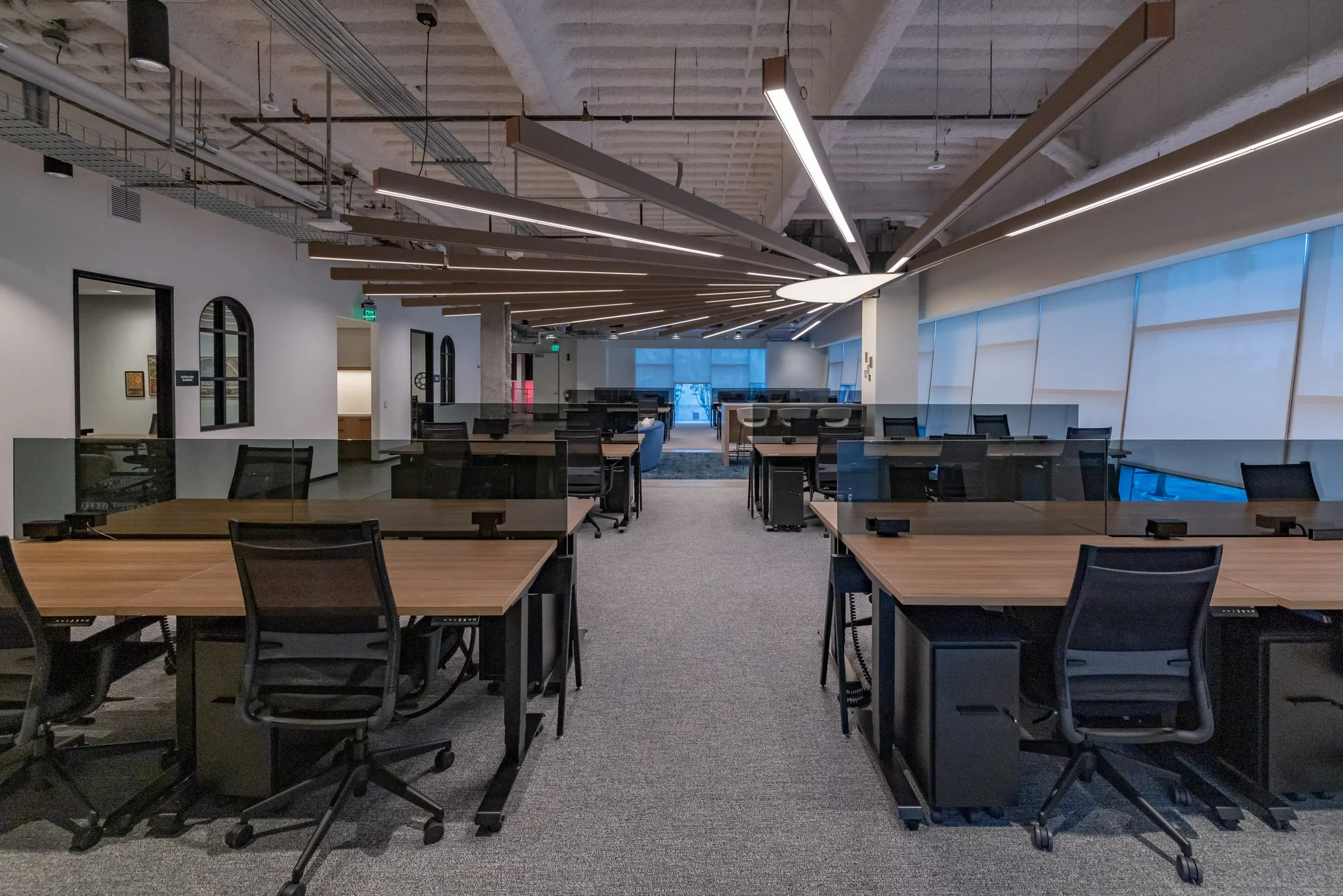 Modern office space with multiple workstations, black chairs, glass partitions, and contemporary ceiling lighting.