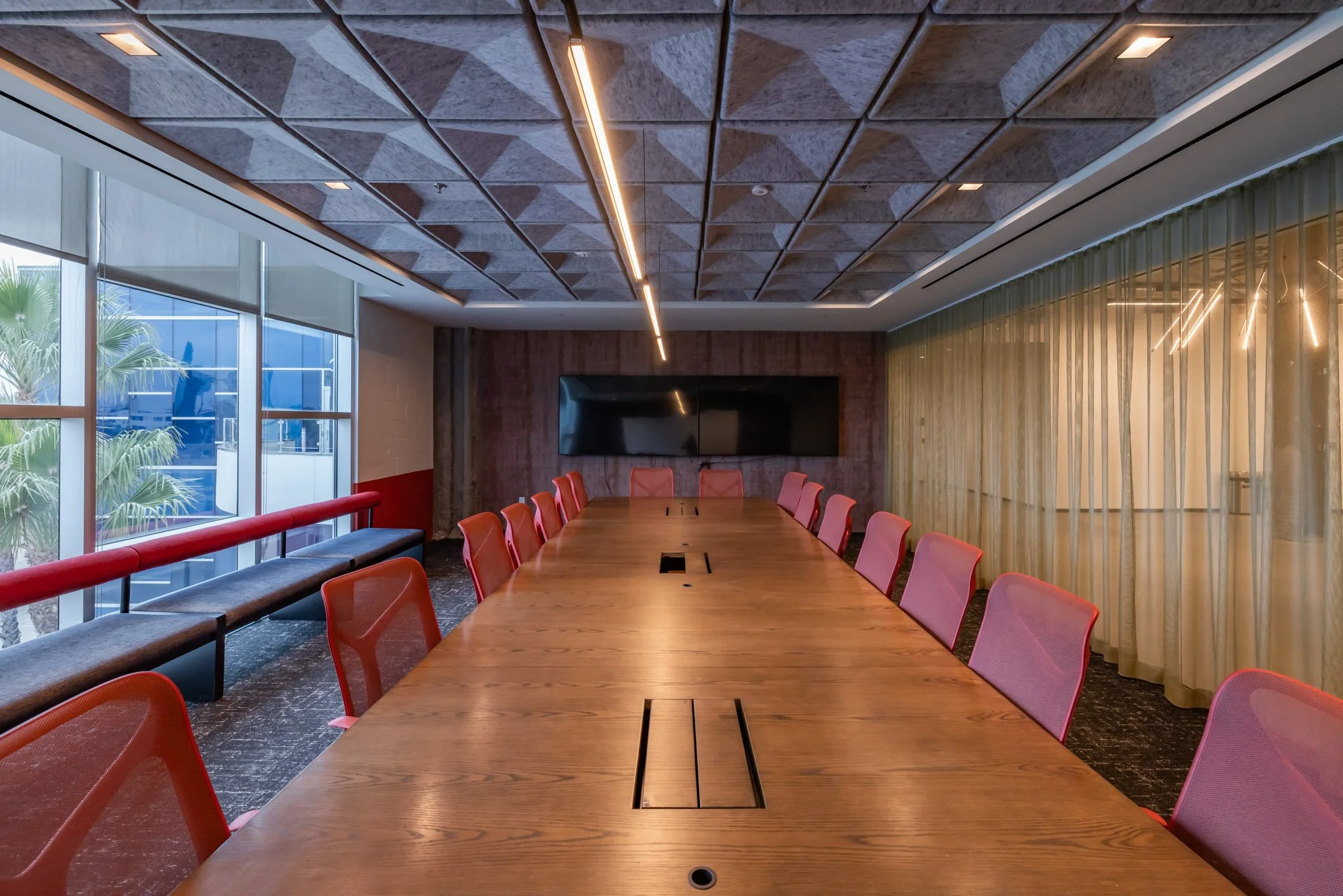 Empty conference room with a long wooden table and pink chairs, large windows on the left, a wall-mounted screen, and modern ceiling lighting.