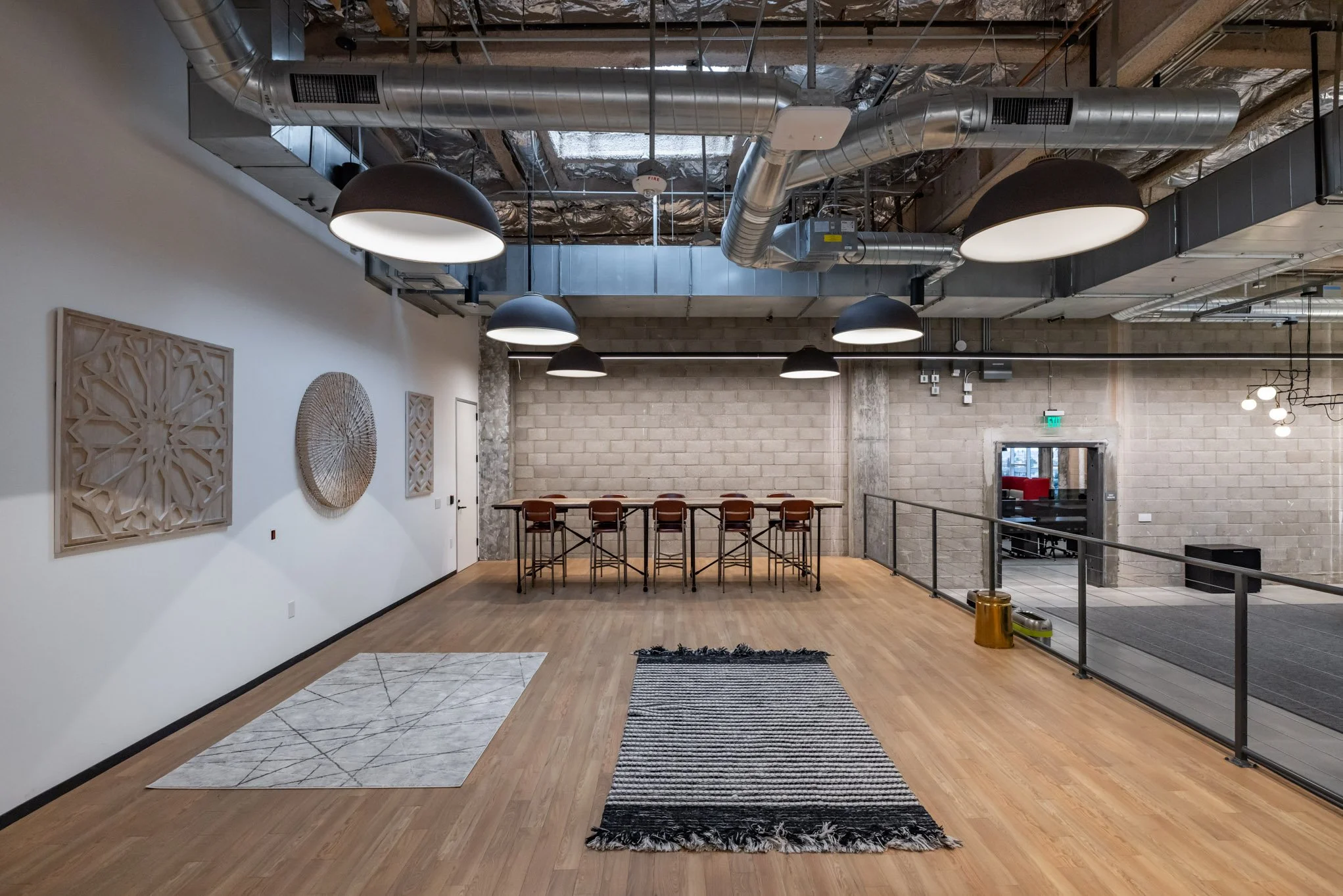 Modern, empty office or coworking space with wooden floors, abstract wall art, hanging black and white pendant lights, high ceiling with exposed ducts, a table with bar stools, and a small entrance area.