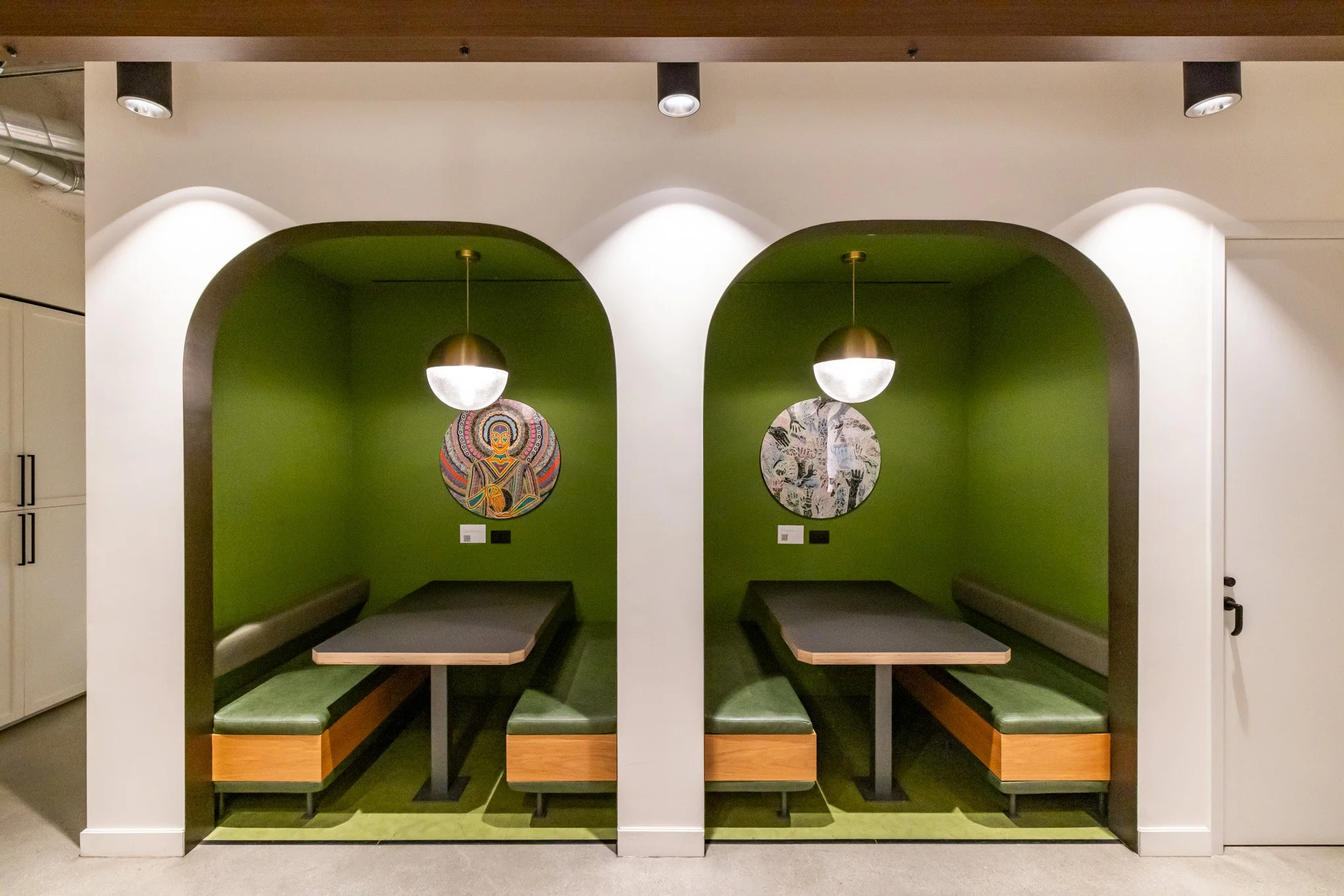 Two built-in booths with green walls, each with a modern pendant light, artwork, and seating, separated by a white wall, in a dining area.