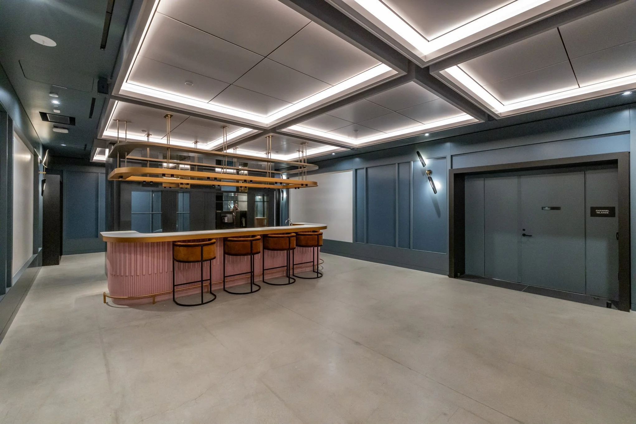 Modern bar with pink curved counter, four brown barstools, overhead wooden structure, blue paneled walls, and sleek ceiling lighting.