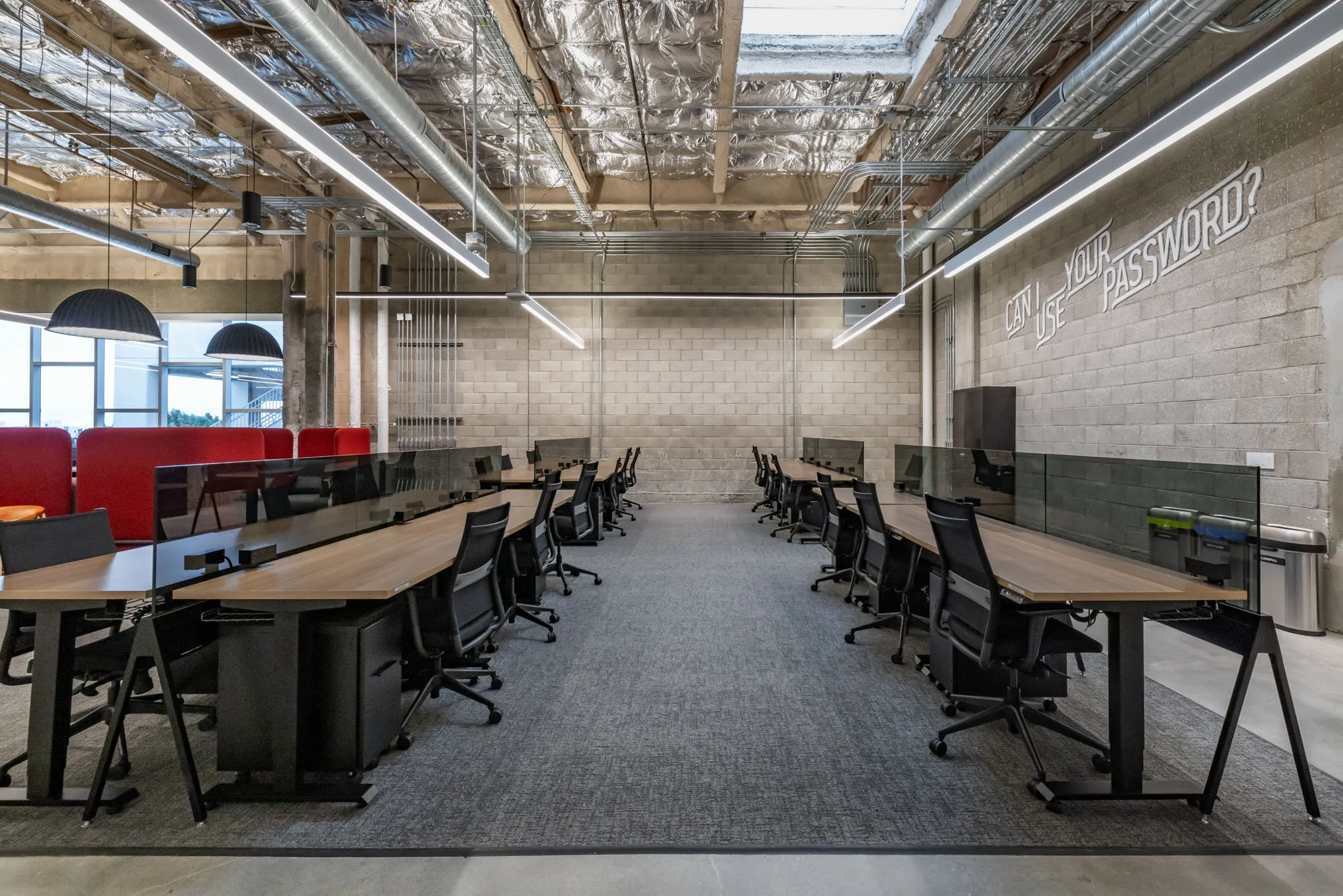 An empty modern conference room with two rows of black office chairs and long wooden tables separated by glass dividers. The room has exposed ductwork and a concrete ceiling with hanging lights, large windows on the left, and a message on the right w