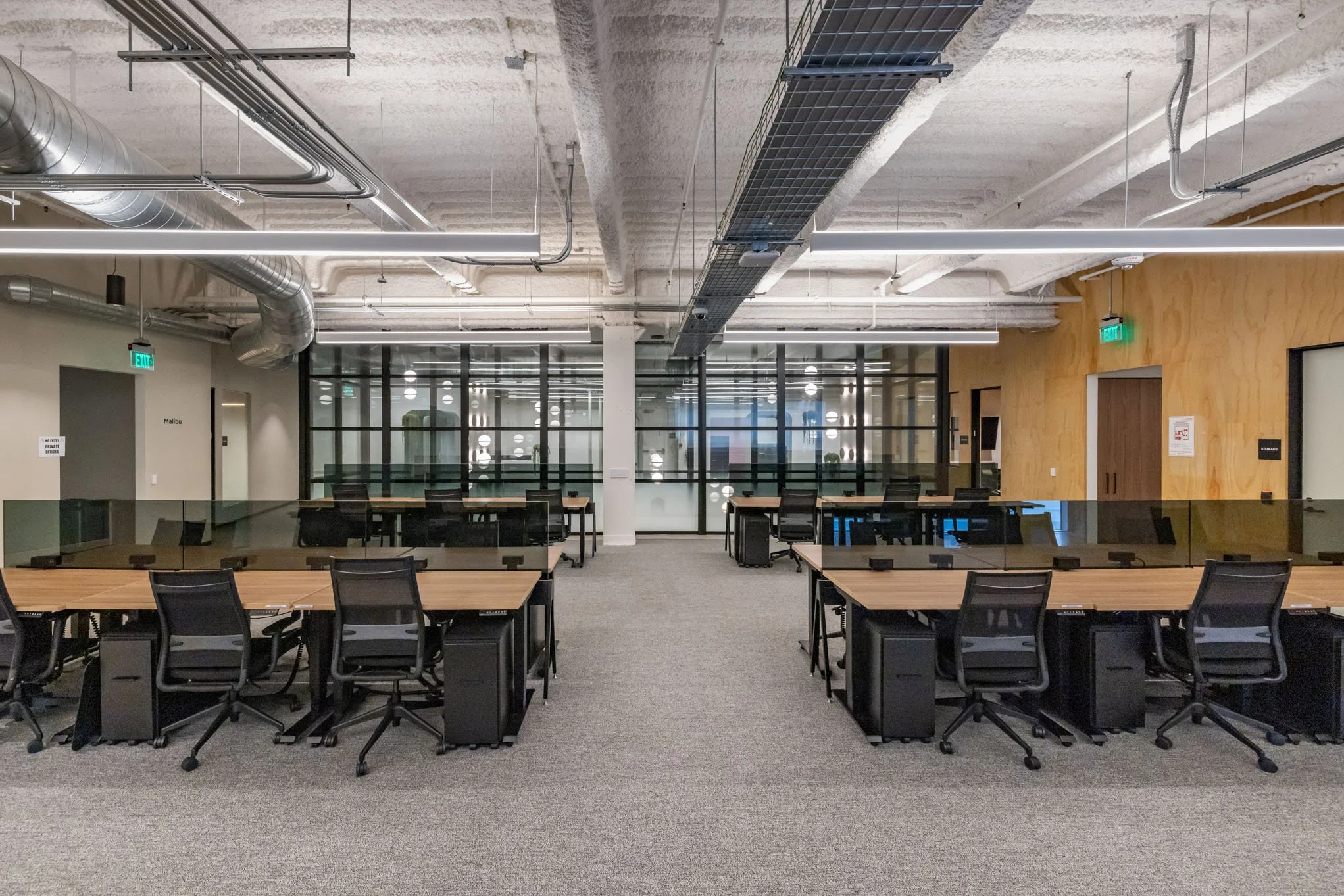 Modern office workspace with multiple desks and chairs, glass-walled meeting rooms, exposed ceiling pipes, and a gray carpeted floor.