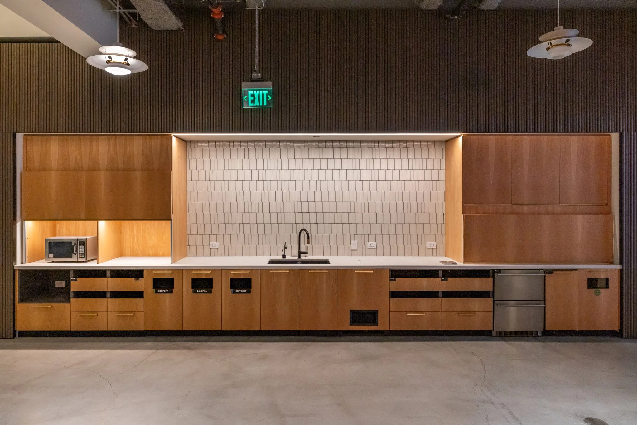 Kitchen area with wooden cabinets, white countertop, black sink, white tiled backsplash, microwave on the left, and various labeled recycling bins beneath the counter. There is a dark wooden wall above and ceiling lights.