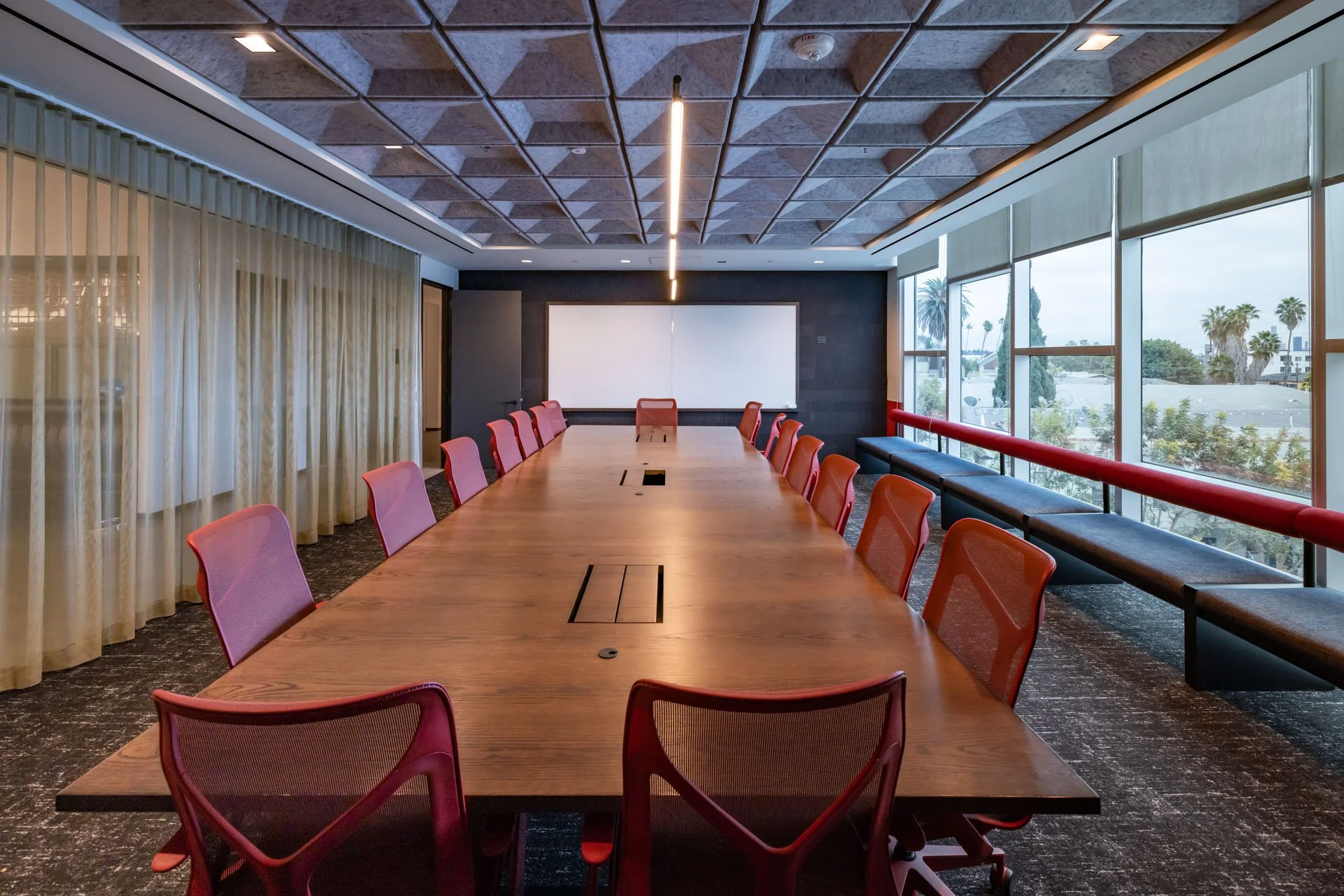 Empty conference room with a long wooden table, red chairs, large windows with a view of palm trees, a whiteboard at the front, and ceiling and wall paneling.
