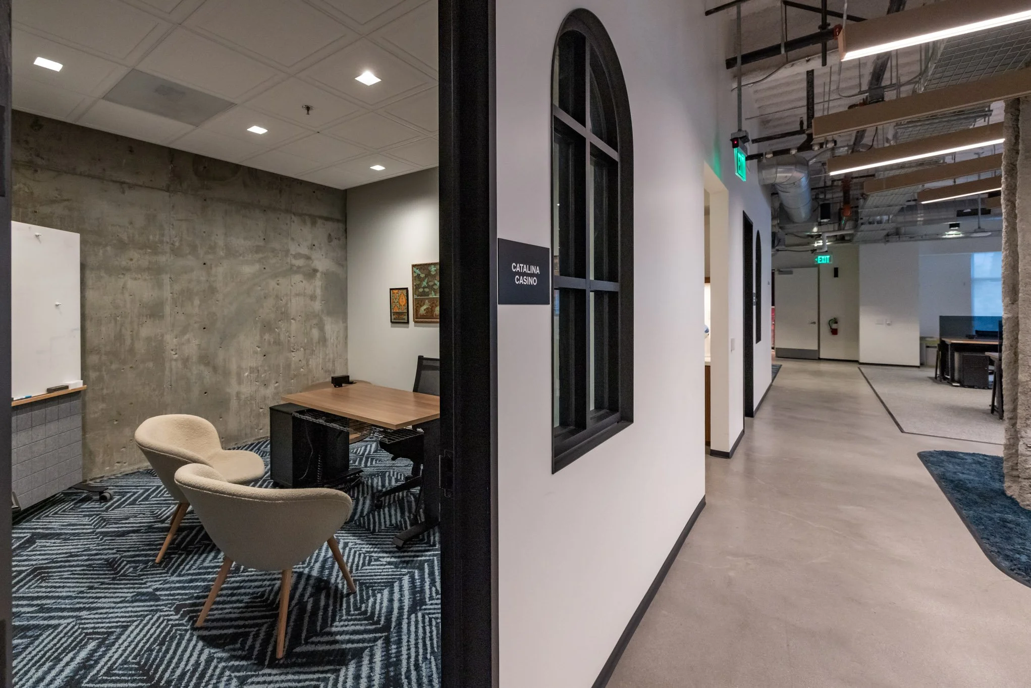 Office space with a conference room labeled 'Catalina Casino' on the door, small table and two beige chairs inside, modern exposed ceiling with ducts, exit signs, and hallway leading to other rooms.