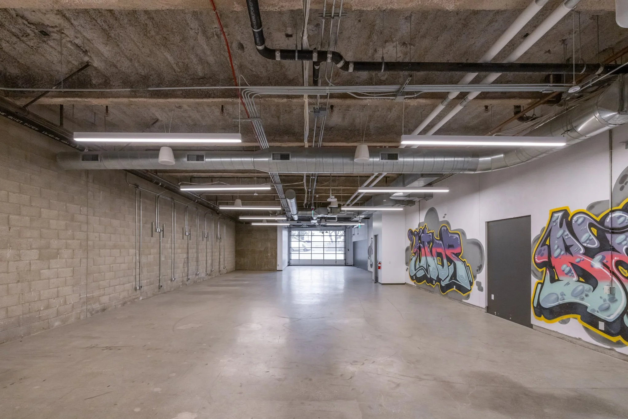 Empty industrial-style parking garage with graffiti art on the right wall, exposed brick and concrete walls, overhead fluorescent lighting, and visible ductwork and piping on the ceiling.