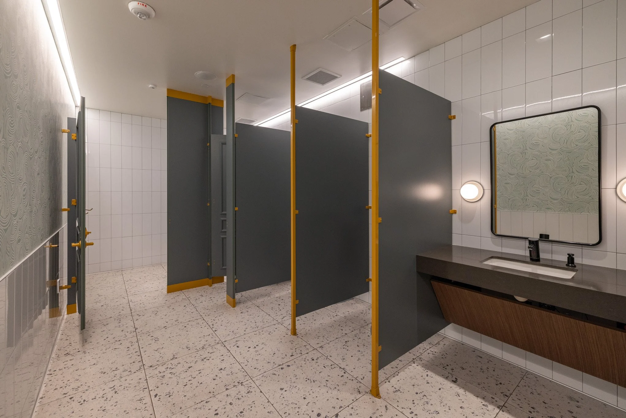 Public restroom with grey and yellow divider stalls, white tiled walls, a sink with a mirror, and wall-mounted lights.