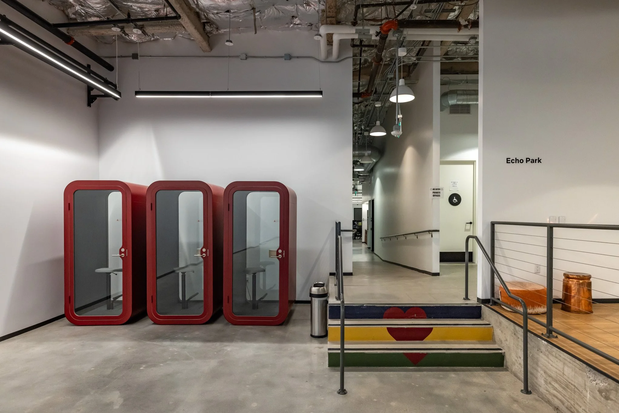 Three red phone booths with glass doors are lined up against a white wall inside a modern building. Next to them are the steps painted with a rainbow pattern and a red heart, leading to a hallway marked $Echo Park. To the side, there's a small orange