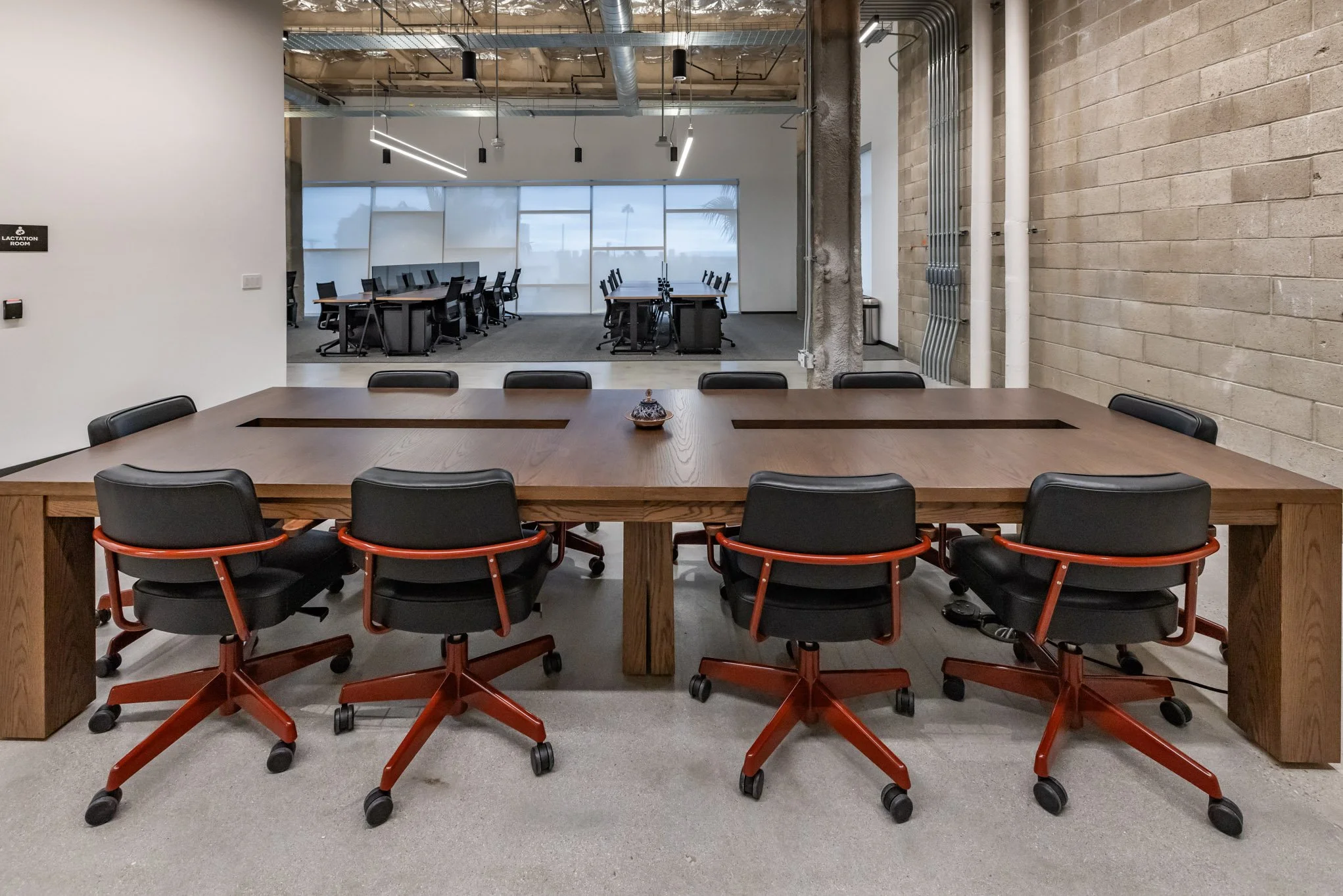 Empty conference room with a large wooden table surrounded by black office chairs with red bases, and a separate larger meeting room with multiple tables and chairs visible through an open partition.