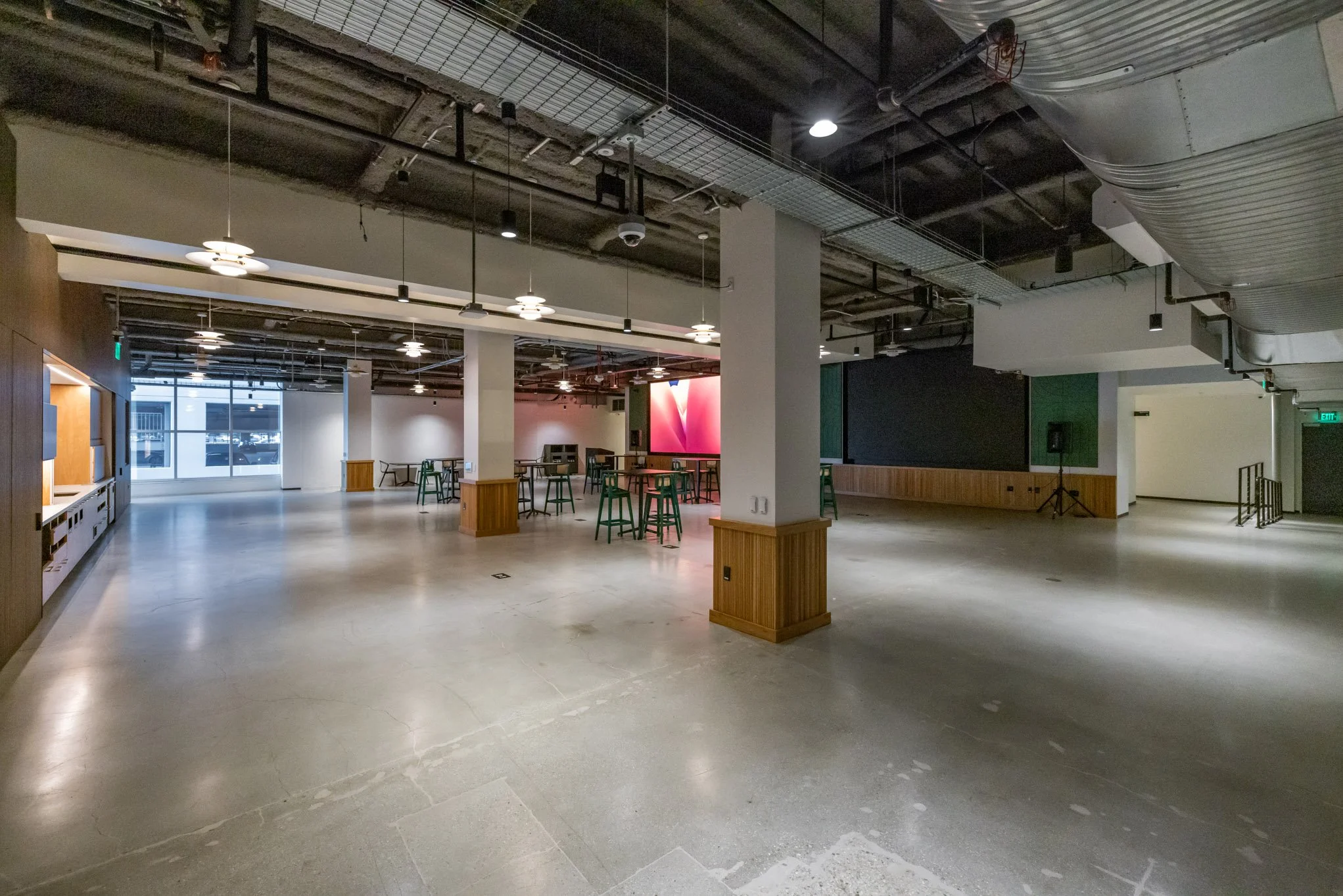Empty event space or lounge area with scattered tables and chairs, large window, carpeted and exposed ceiling with industrial fixtures and lighting.