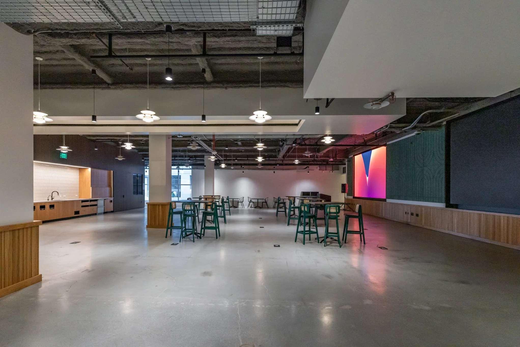 An empty, modern indoor lounge or event space with high ceilings, ceiling lights, and a large digital screen on the wall. There are tall tables with chairs and a kitchenette area to the left.