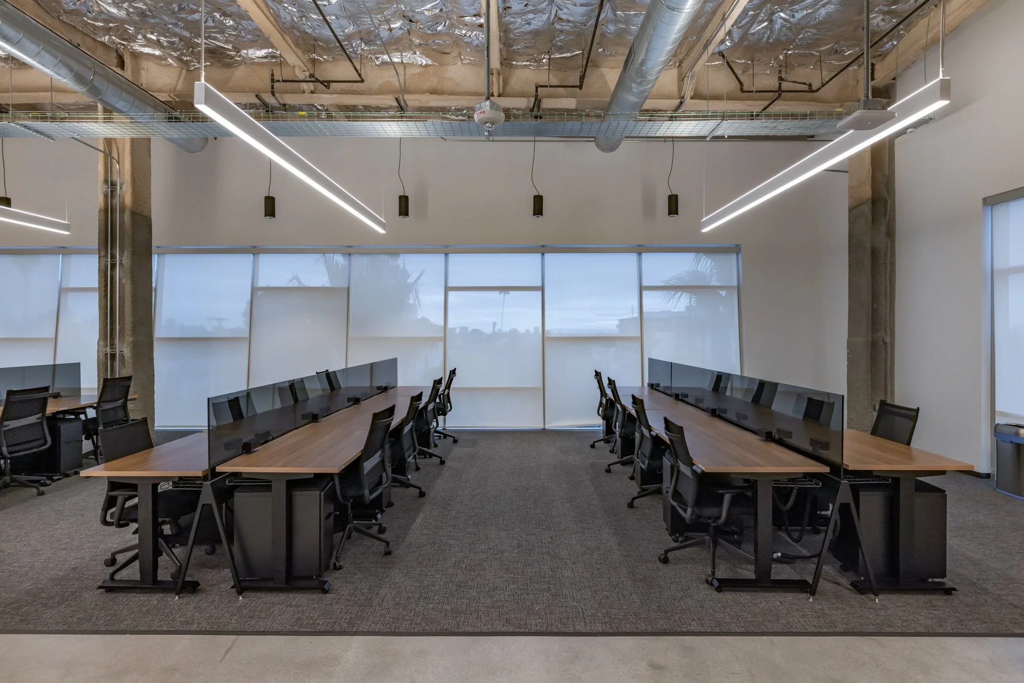 Empty modern conference room with two long tables and black chairs separated by transparent dividers, large windows with roller shades, and exposed ceiling with pipes and lighting fixtures.
