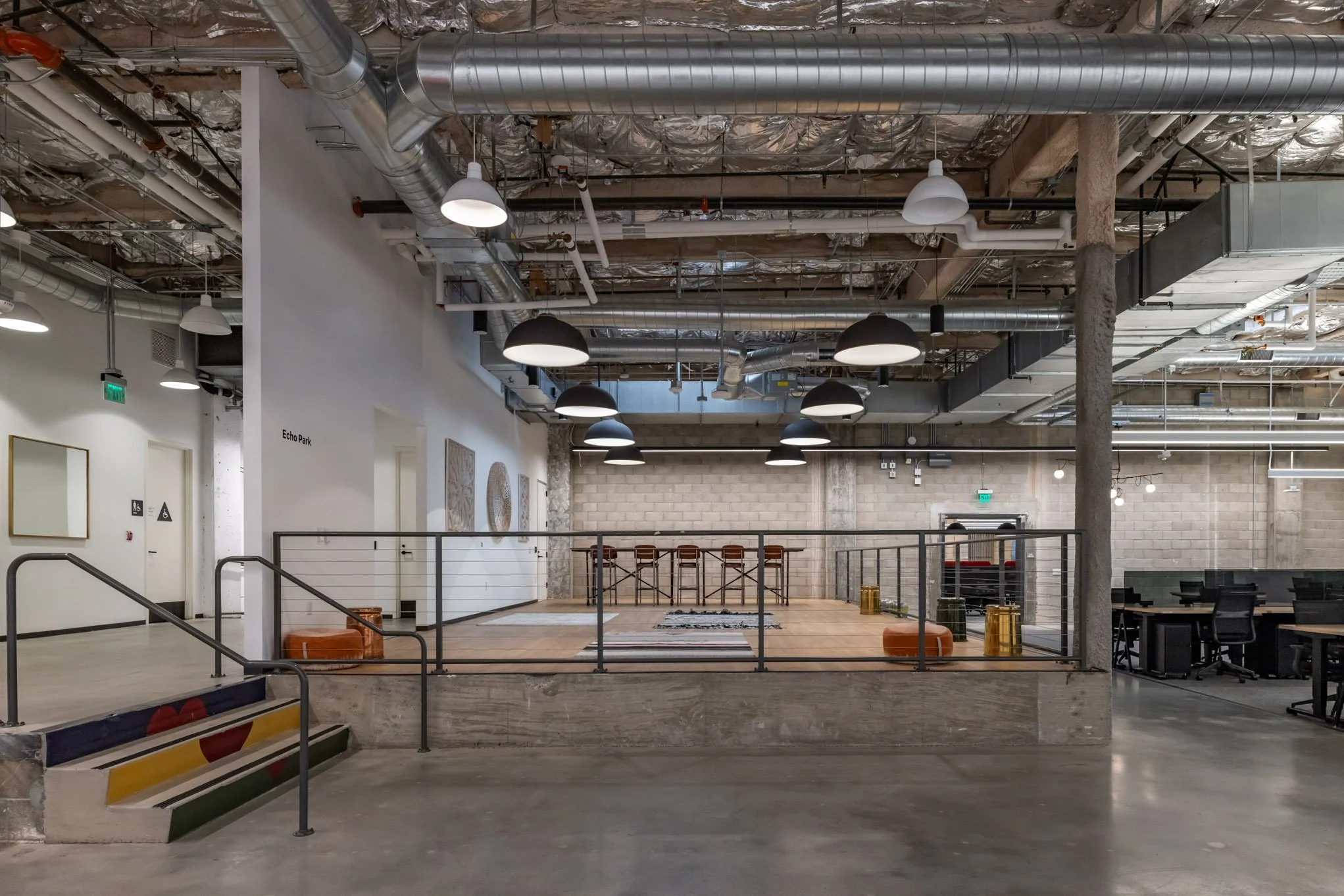 Interior of a modern office space with a small stage or platform, wooden flooring, industrial ceiling with exposed ducts and pipes, black hanging lights, and workstations with black chairs on the right side.