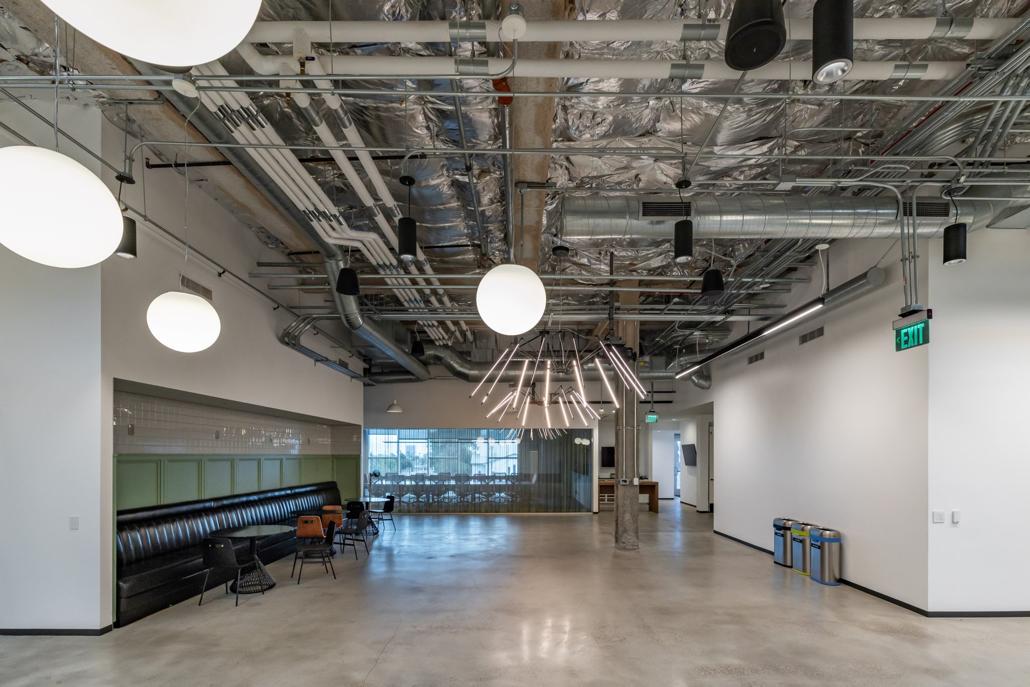 Empty modern lobby or communal space with exposed ceiling pipes and ducts, pendant and LED light fixtures, a black leather bench, small chairs and tables, large windows with blinds, and an exit sign.