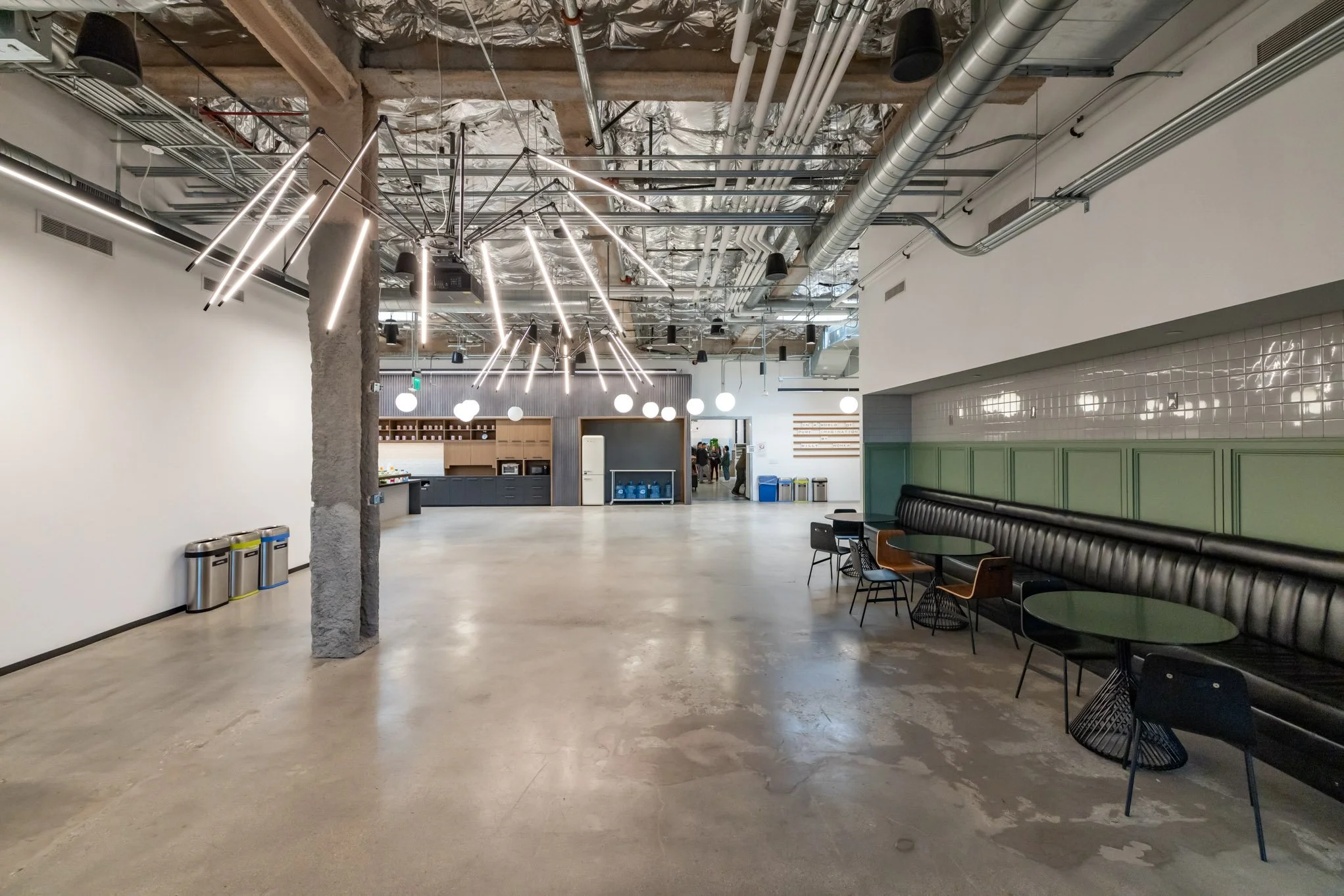 A modern, open-concept lobby with polished concrete floors, white walls, and an exposed ceiling with ductwork and pipes. There are several black round tables with chairs along a black leather bench against the right wall. A unique chandelier with mul