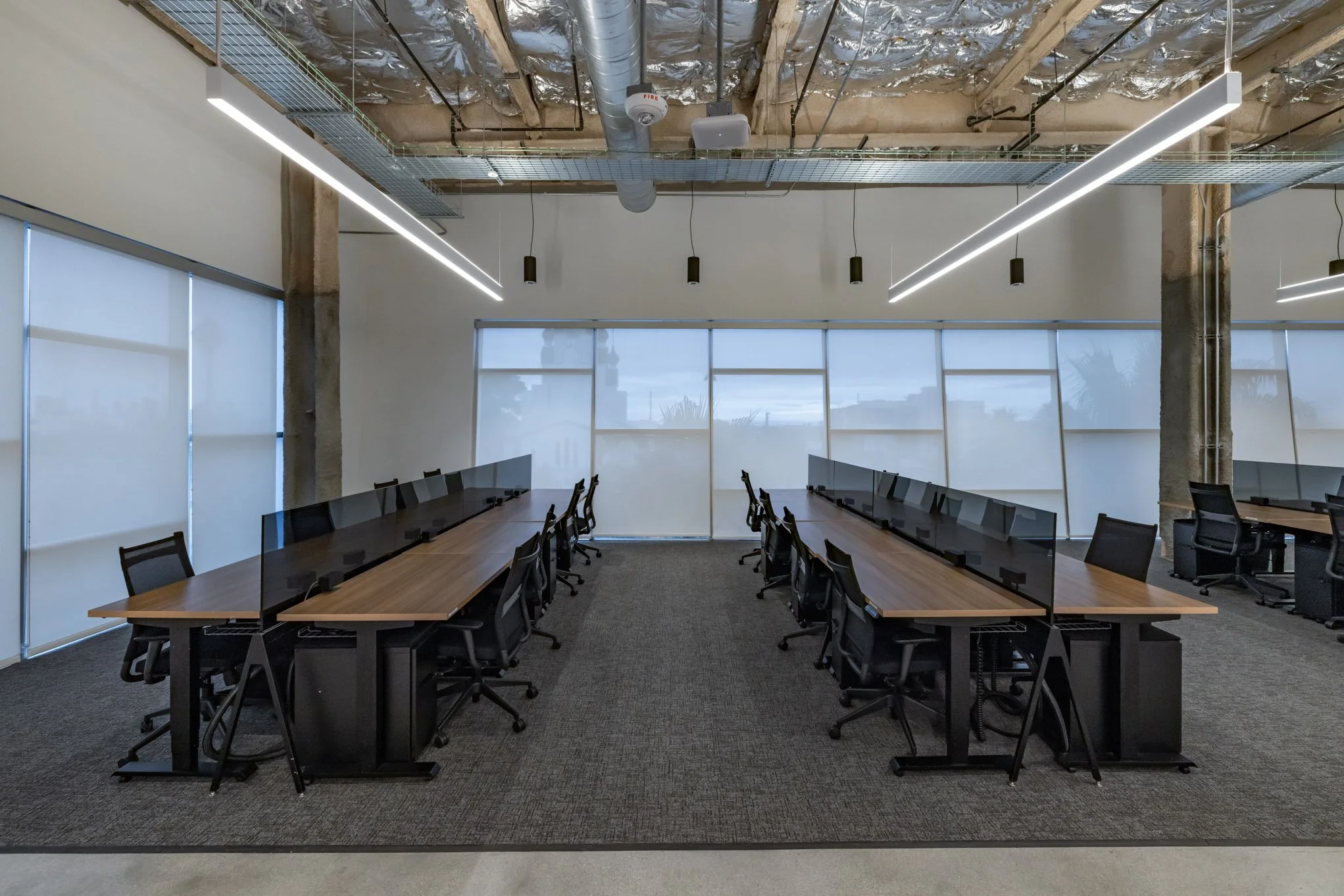 Modern empty office conference room with long wooden tables, black chairs, and glass privacy screens, large windows with blinds, and industrial ceiling with exposed pipes and lighting.