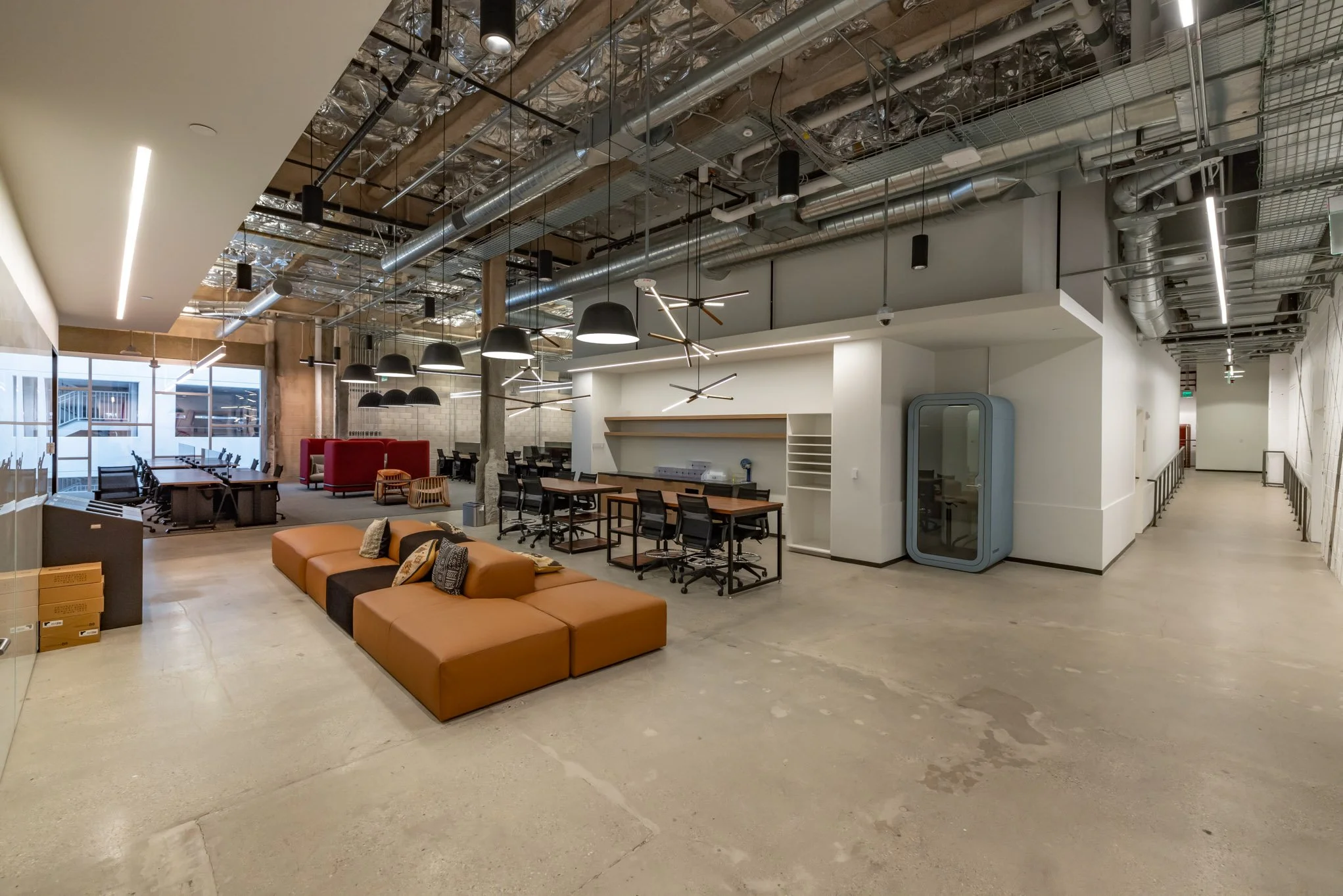 Open office space with modern furniture, black chairs at wood tables, orange seating in foreground, black pendant lights, exposed ceiling with ductwork, large windows, and a hallway to the right.