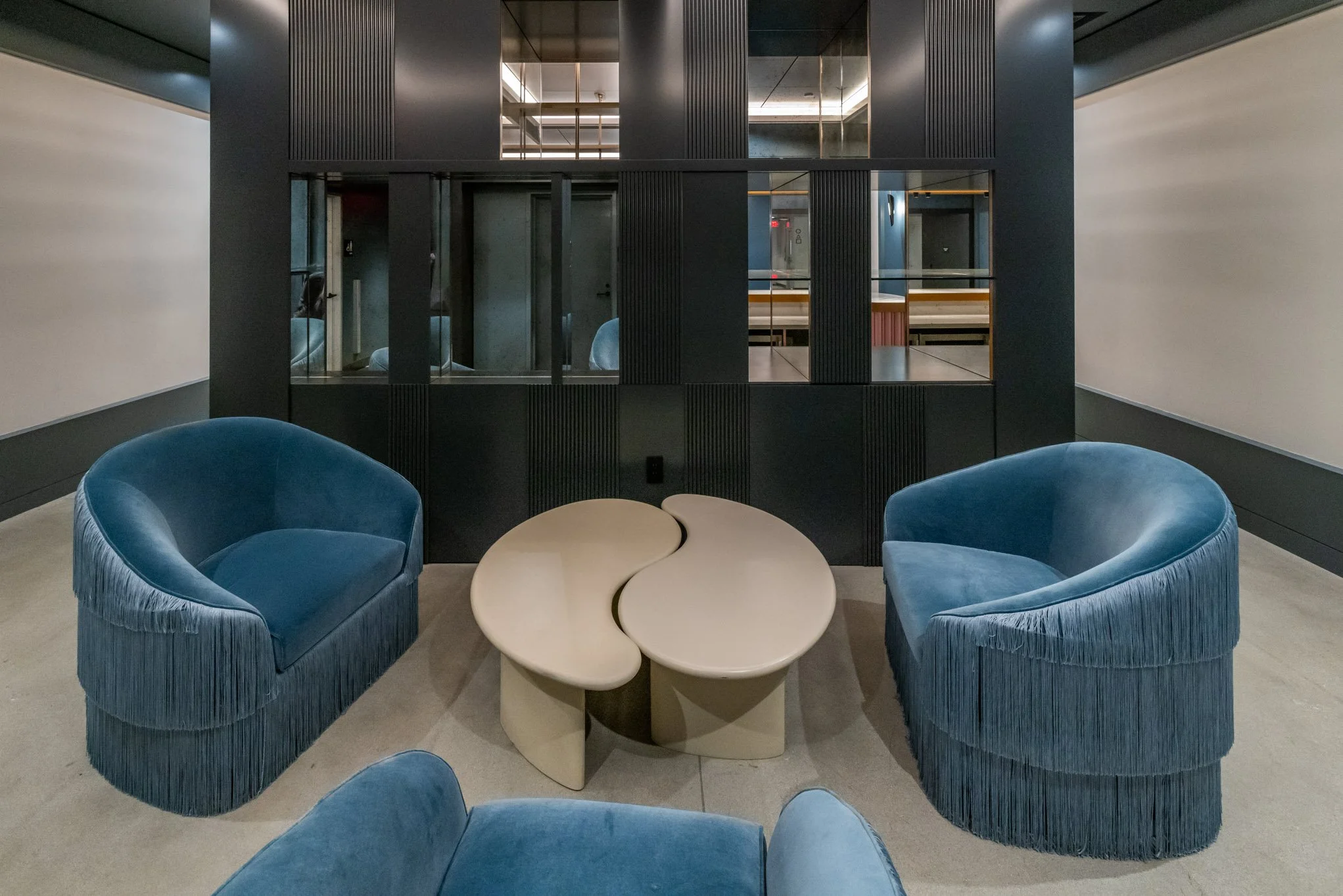 Two blue velvet armchairs with fringe trim facing each other, separated by a beige, abstract, yin-yang-shaped coffee table, in a modern interior with dark walls, reflective black panels, and a mirror wall.