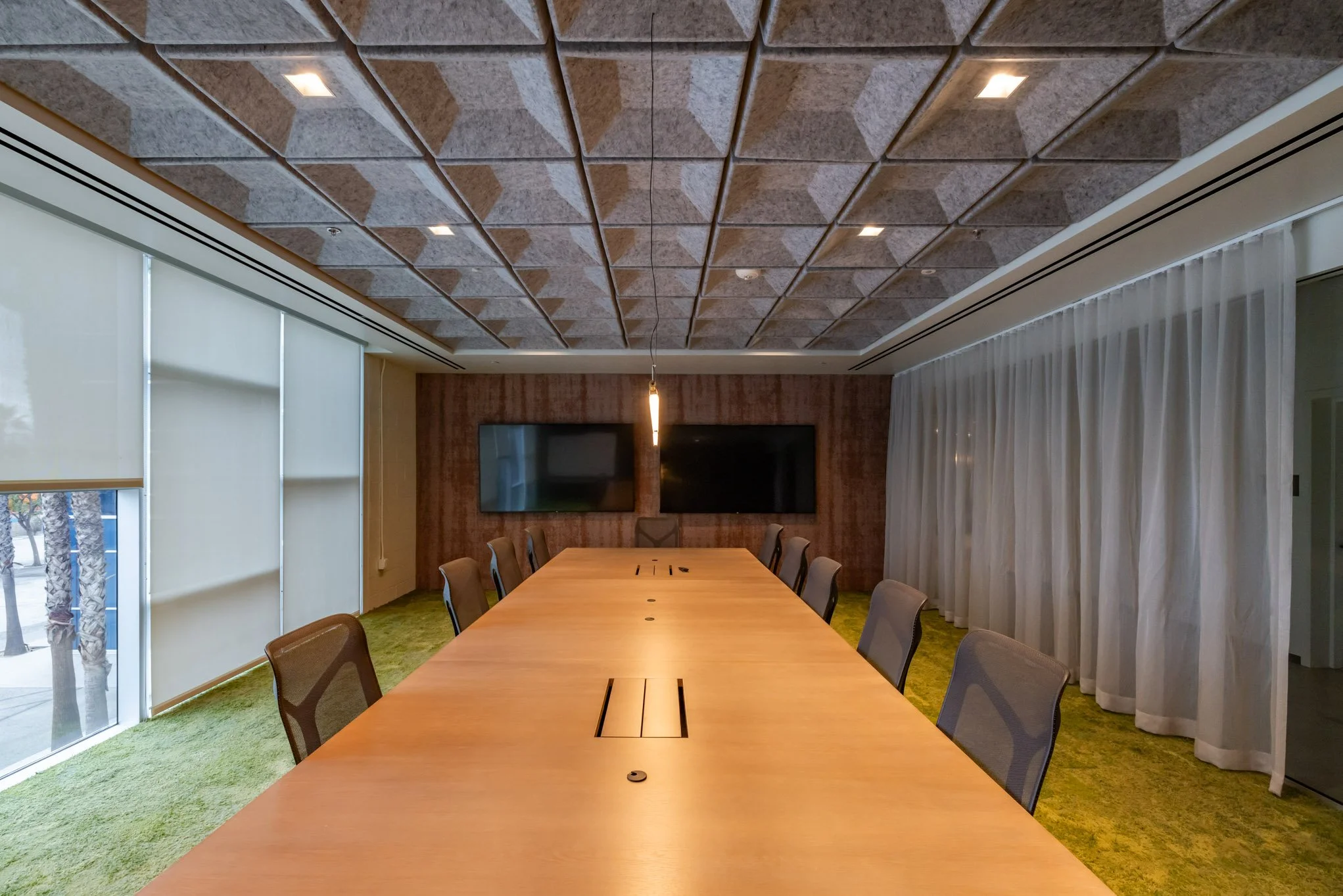 Empty conference room with a long wooden table, chairs, large windows with blinds, a textured ceiling, and two mounted televisions on the back wall.