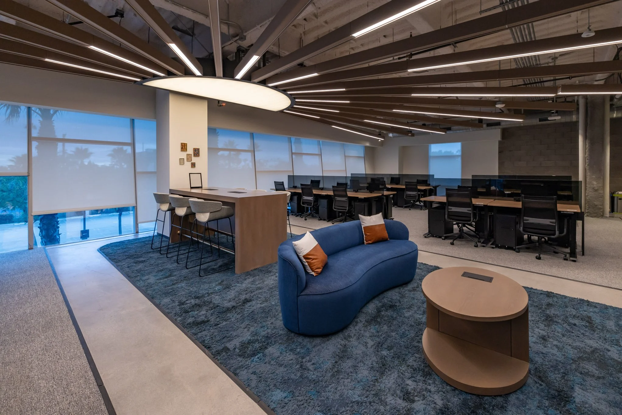 Modern office lounge and workspace with blue sofa, wooden coffee table, bar area with stools, row of desks with office chairs, large windows, and contemporary lighting fixtures.