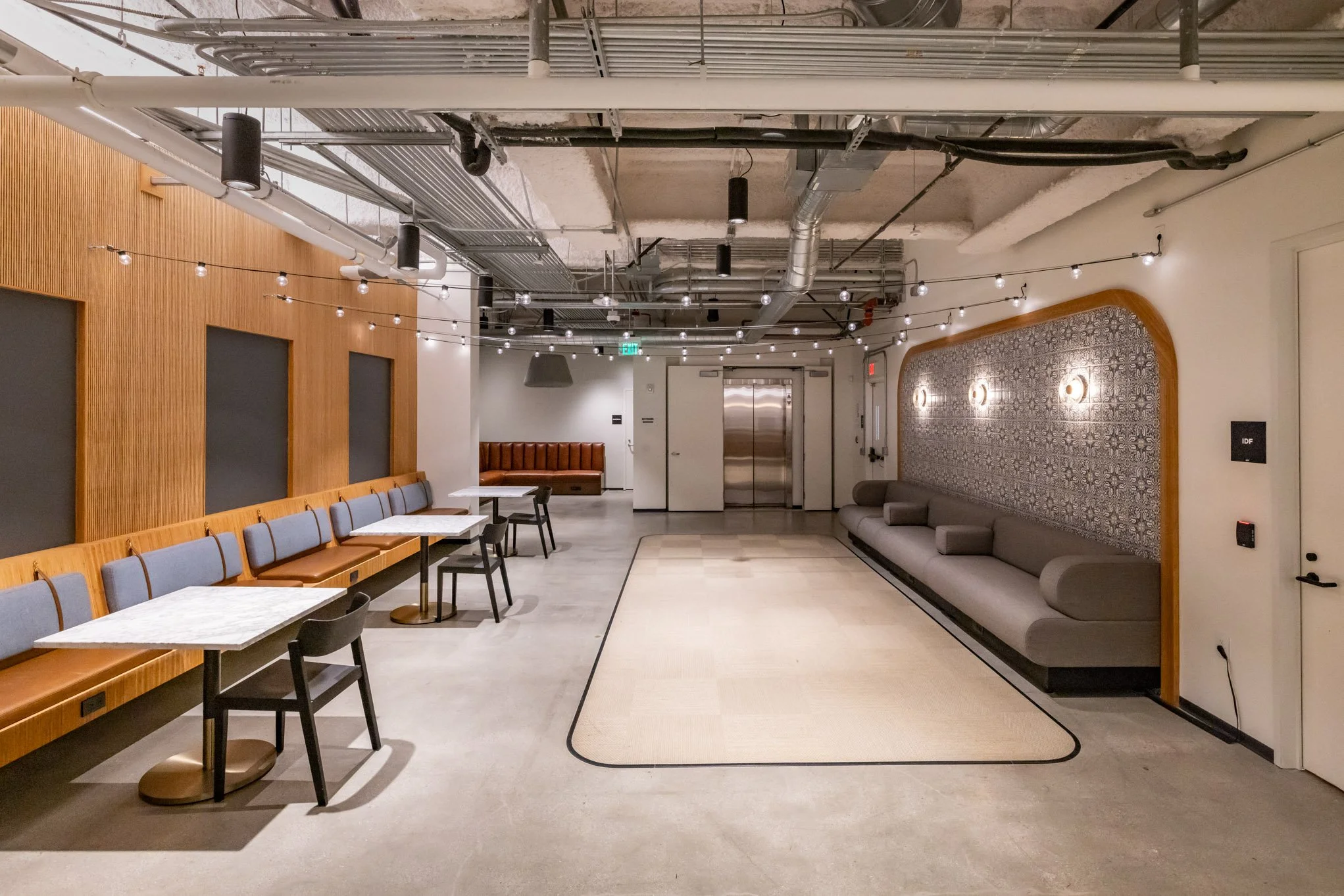 Modern lounge area with blue chairs and white tables on the left, and a long gray sofa on the right, with string lights hanging from the ceiling.