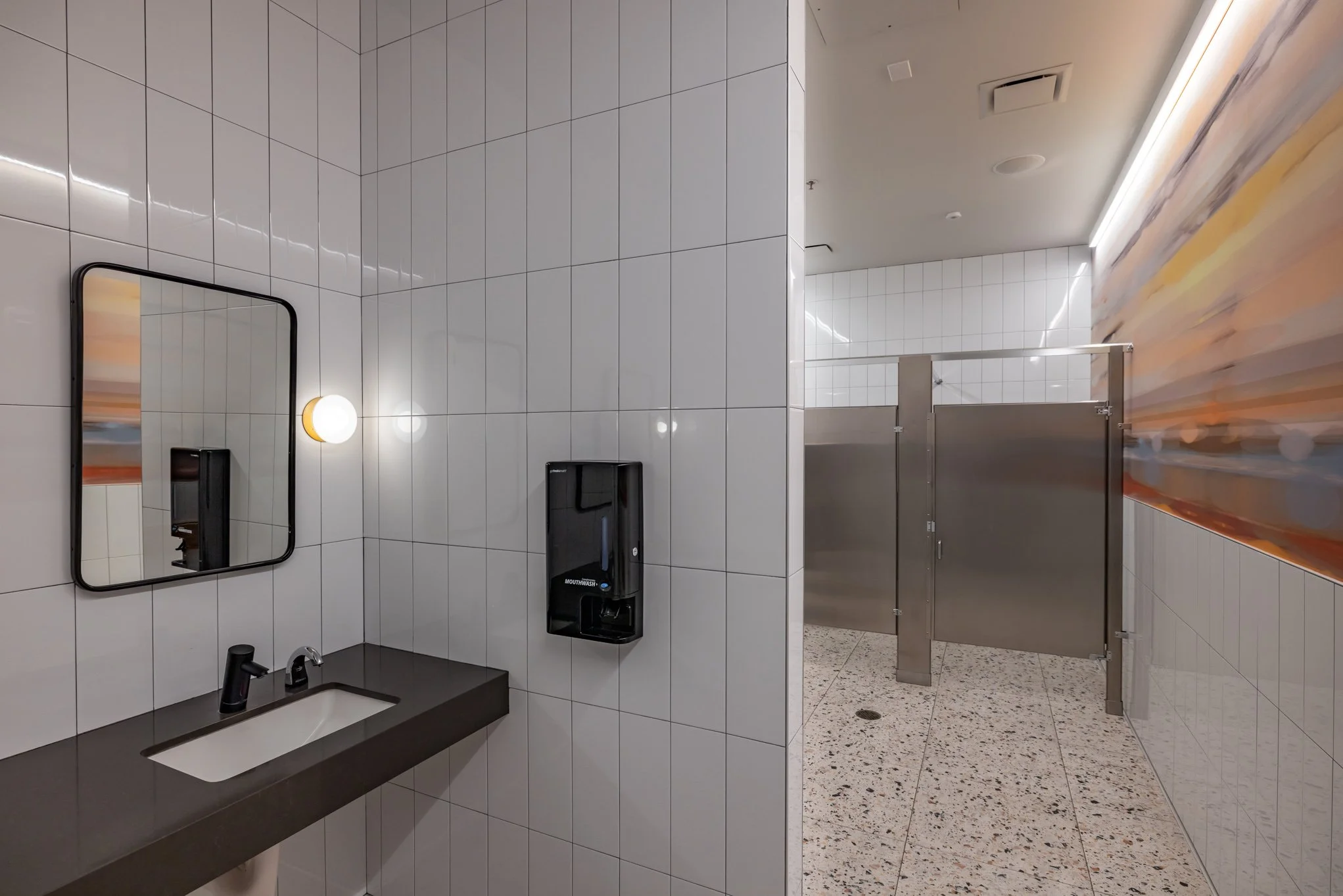 Public bathroom with white tiled walls and a dark countertop sink, mirror, wall light, and a black hand sanitizer dispenser; stainless steel stalls are visible in the background.