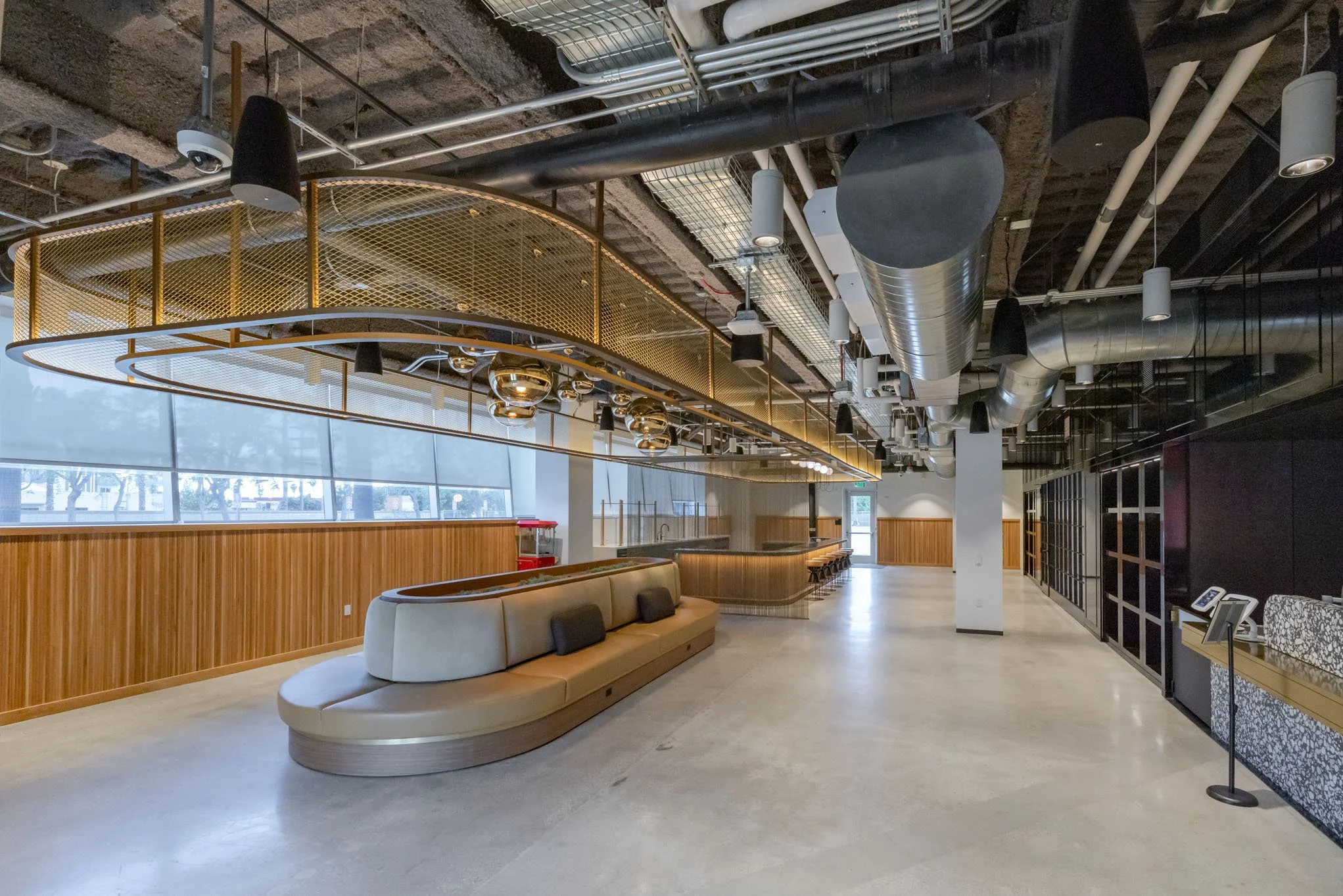 Empty modern interior space with a curved beige seating area, wooden wall paneling, large windows, and an open ceiling with exposed ductwork and pipes.