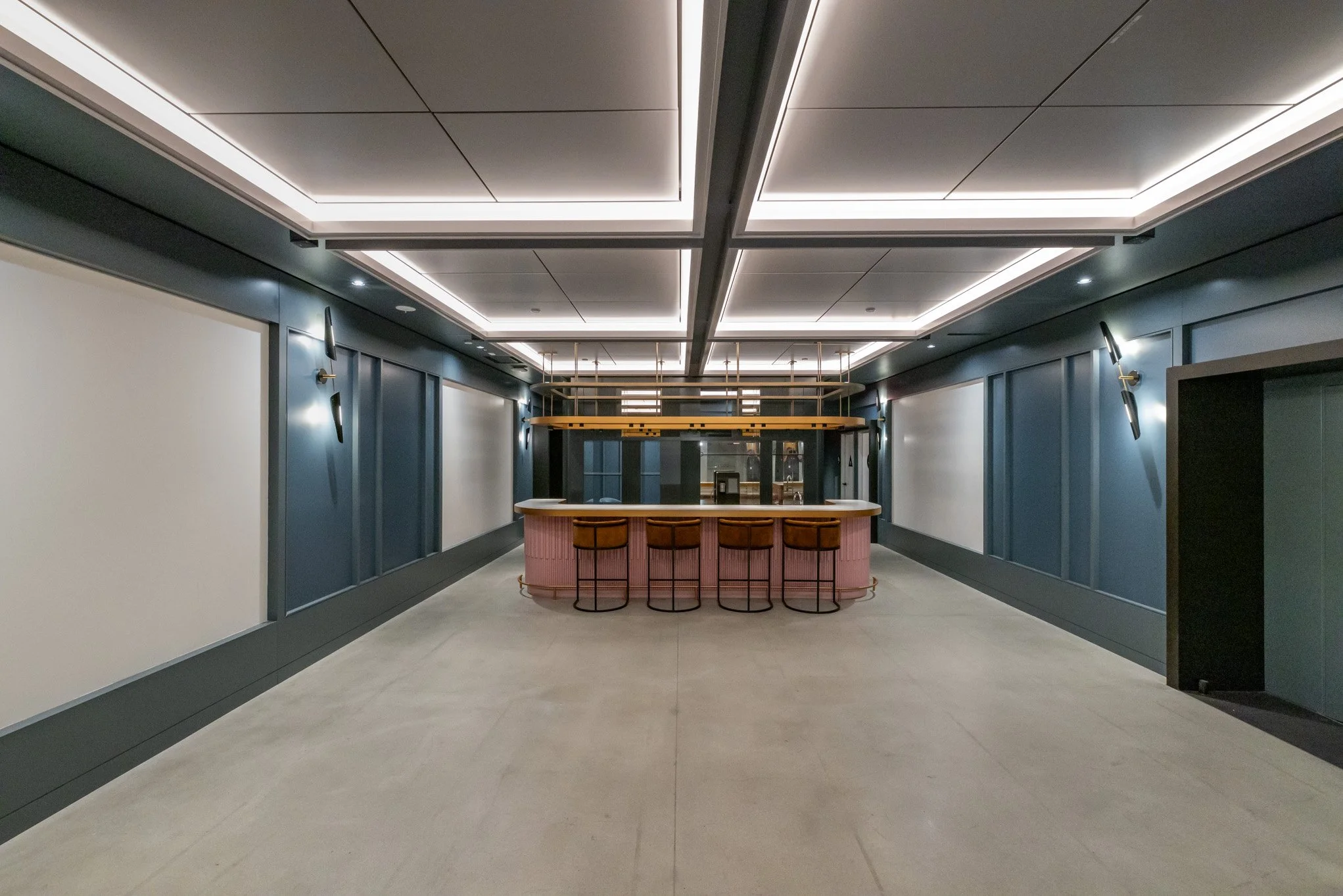 Empty modern lounge area with a circular pink bar counter in the center and four brown bar stools. Blue-paneled walls with wall sconces, overhead lighting, and a glass wall in the background.