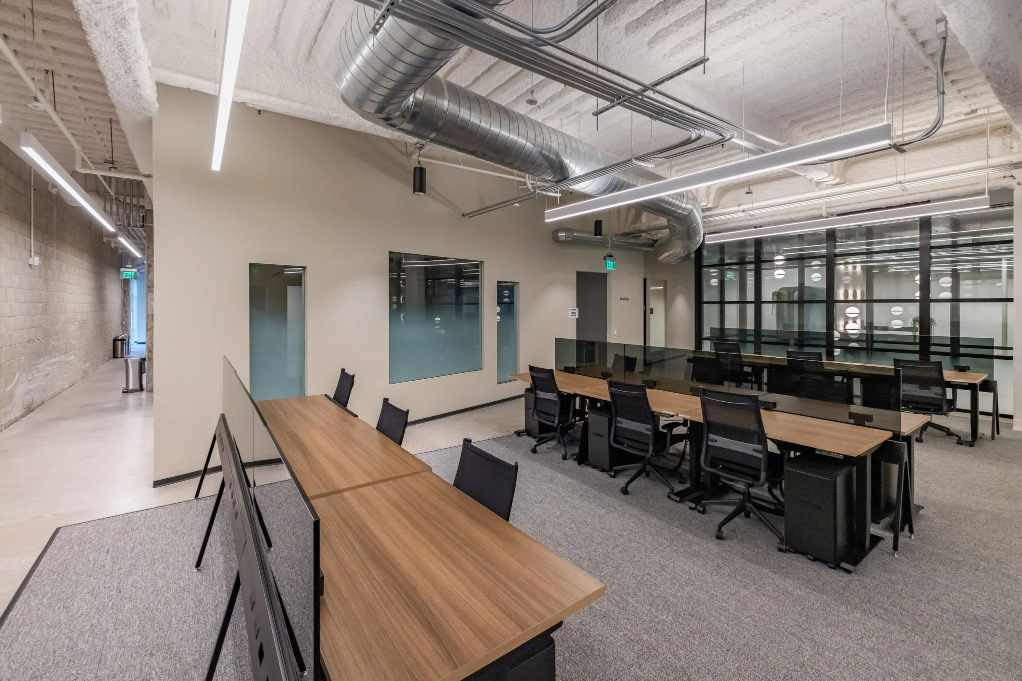 Modern office space with wooden desks, black chairs, glass partitions, exposed ductwork, and contemporary lighting.