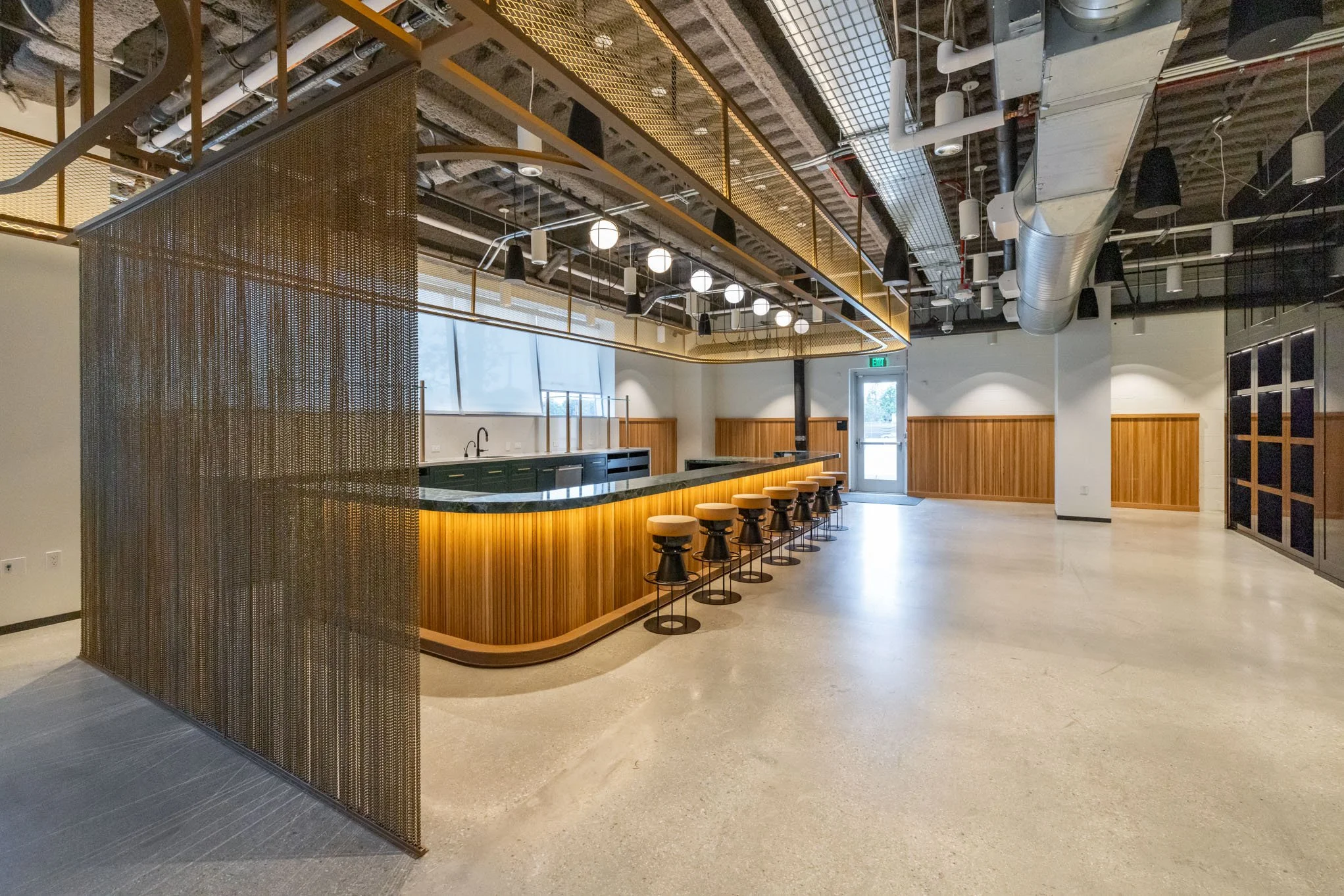 Modern interior space with a curved wooden bar with seven beige barstools, black bases, and a black countertop. Overhead lighting includes dome-shaped and round globe fixtures. The ceiling exposes ductwork and piping, and there is a metal mesh divide