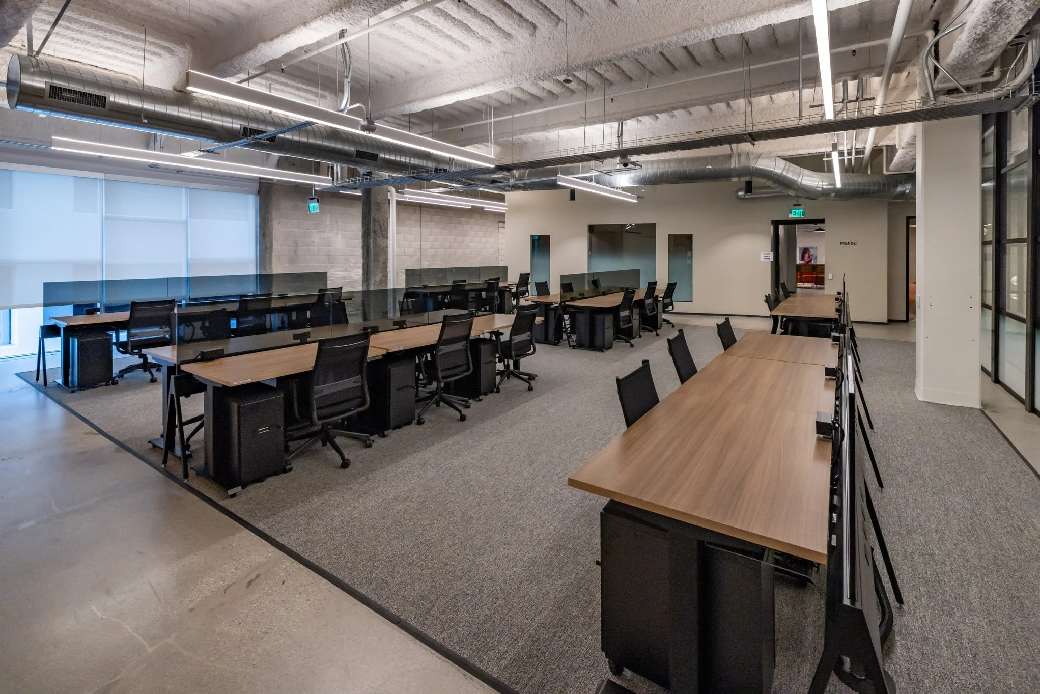 Empty modern office conference room with wooden desks, black chairs, glass dividers, and exposed ceiling ducts.