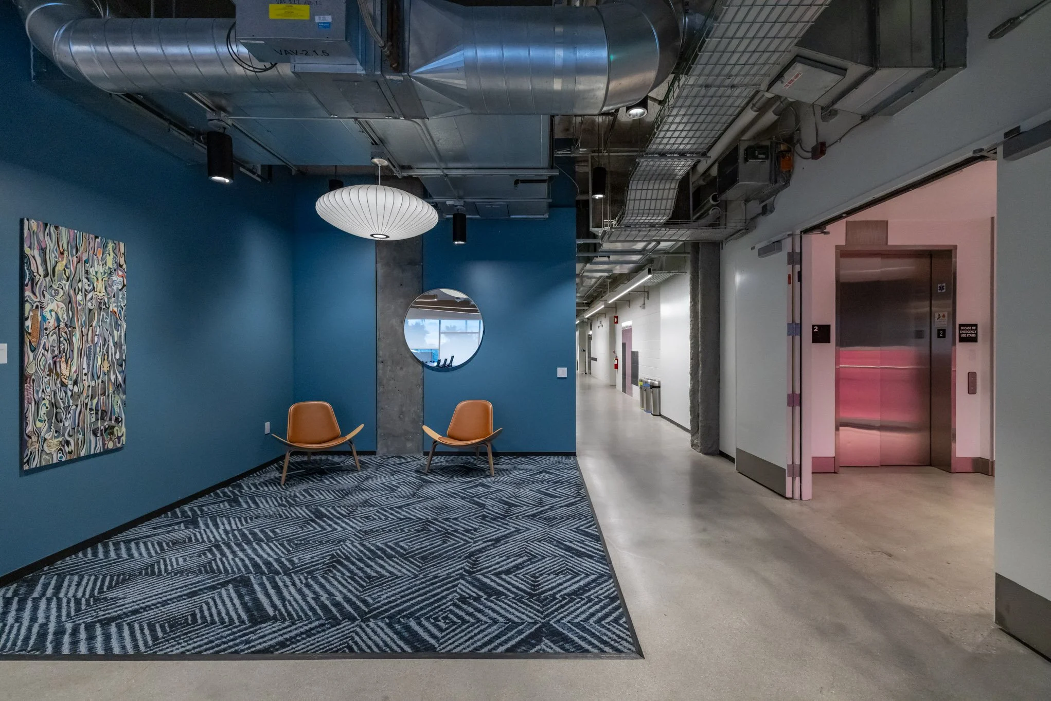 Modern hallway with blue accent wall and seating area featuring two brown chairs, a circular mirror, modern artwork, and an abstract patterned rug. To the right, an elevator with open doors showing pink lighting. The ceiling has exposed ducts and ind