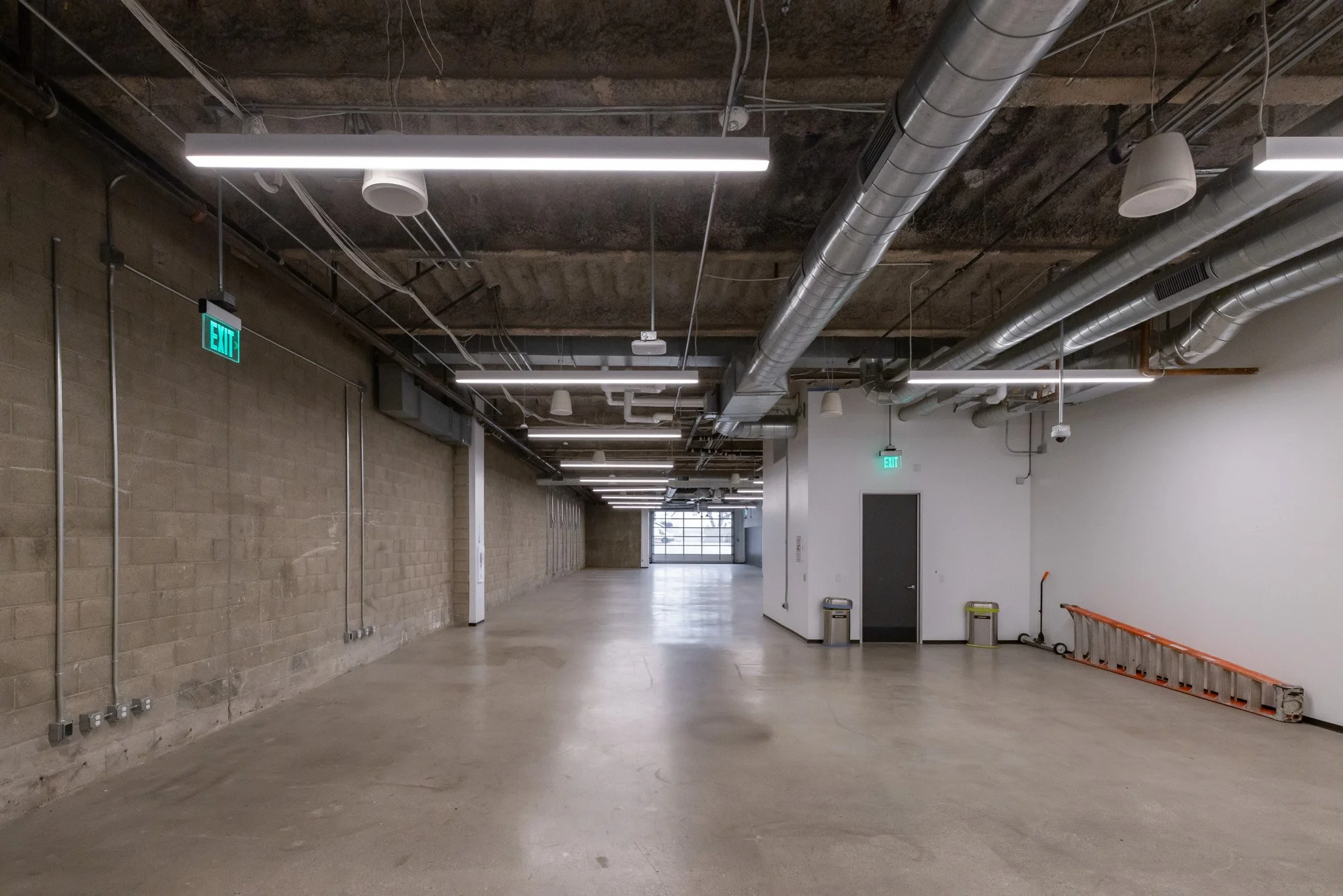 Empty industrial-style indoor space with concrete floor, cinder block wall on the left, white wall on the right, exposed ductwork and piping on ceiling, fluorescent lighting, exit signs, and a metal ladder on the right side.