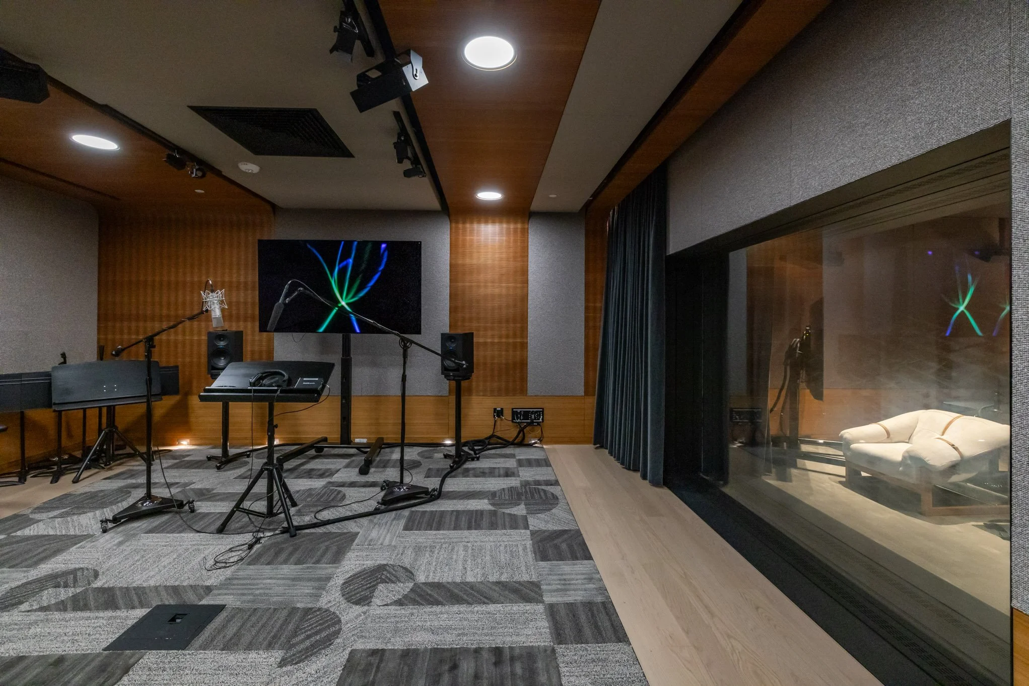 Recording studio with microphones, music stands, speakers, a large screen displaying colorful lines, and a window showing a couch and additional equipment.
