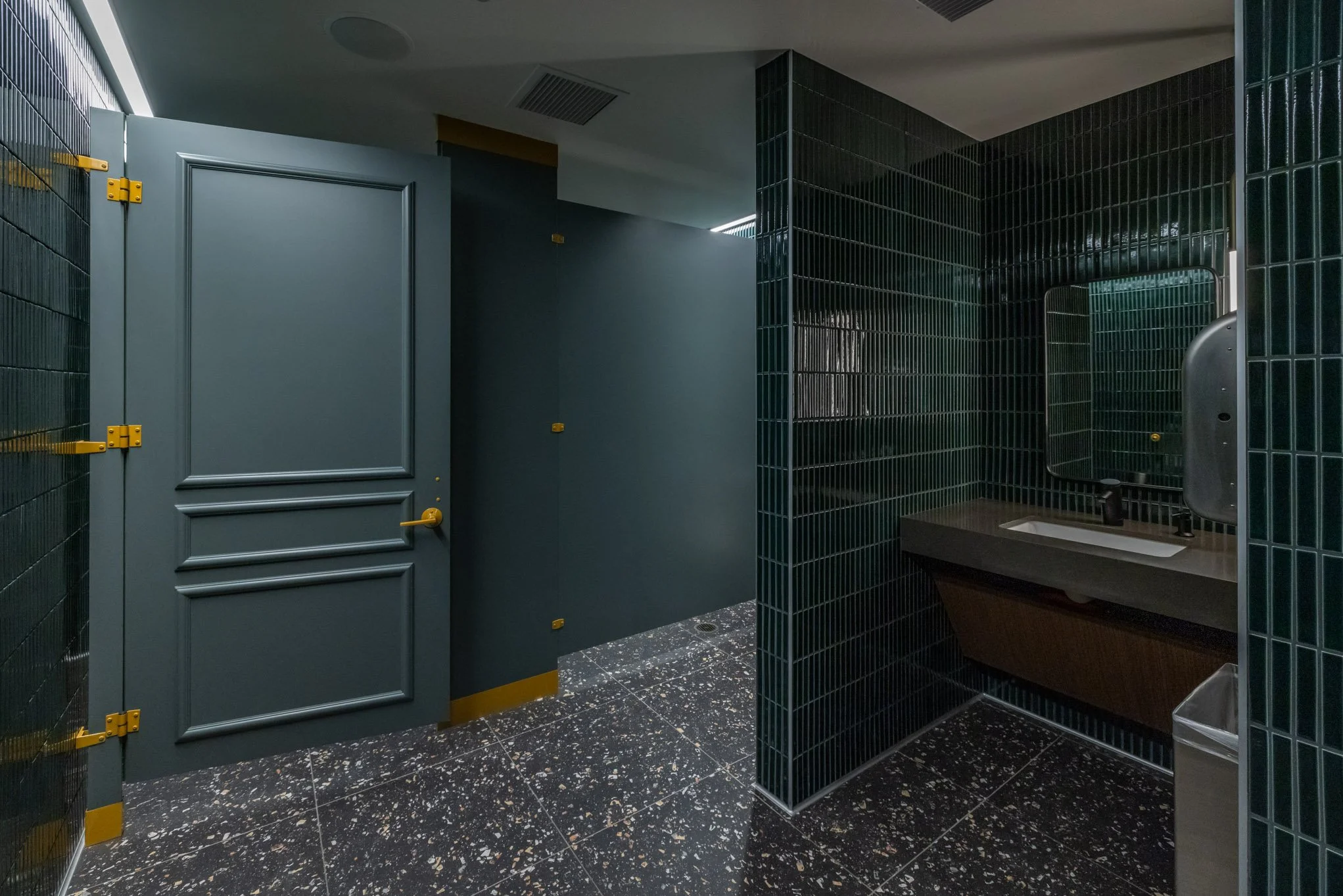 A modern bathroom with dark green tiled walls, a small sink with a black faucet, a large mirror, and a closed blue door. The floor is black with white specks.