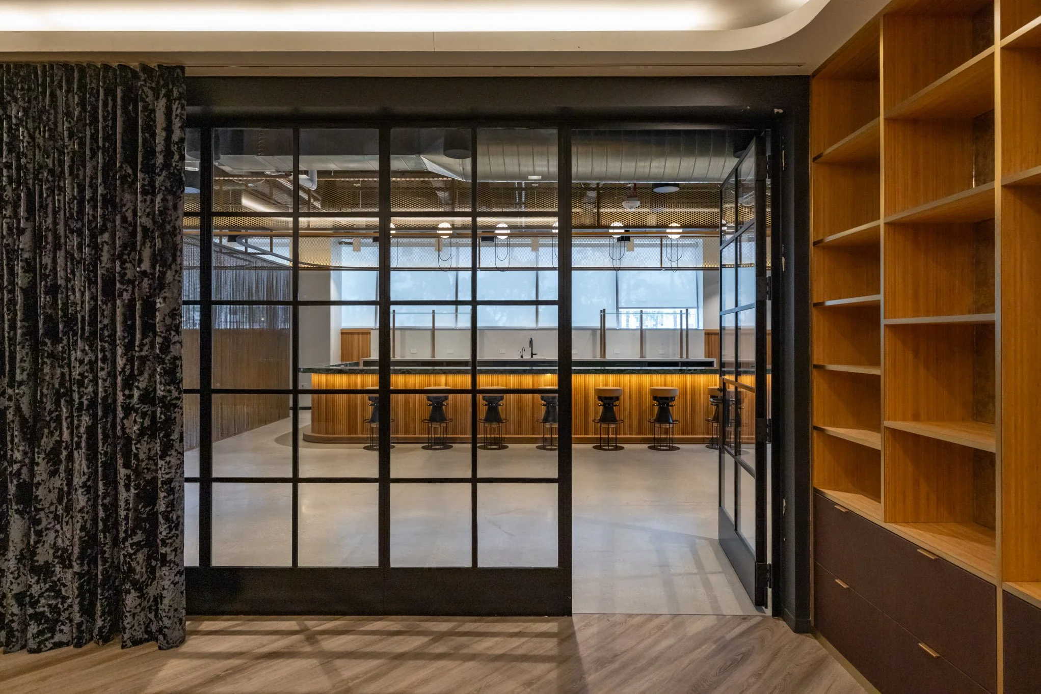 Modern bar with wooden counter and black stools, viewed through black metal framed glass doors, inside a stylish interior space with wooden and patterned curtains.