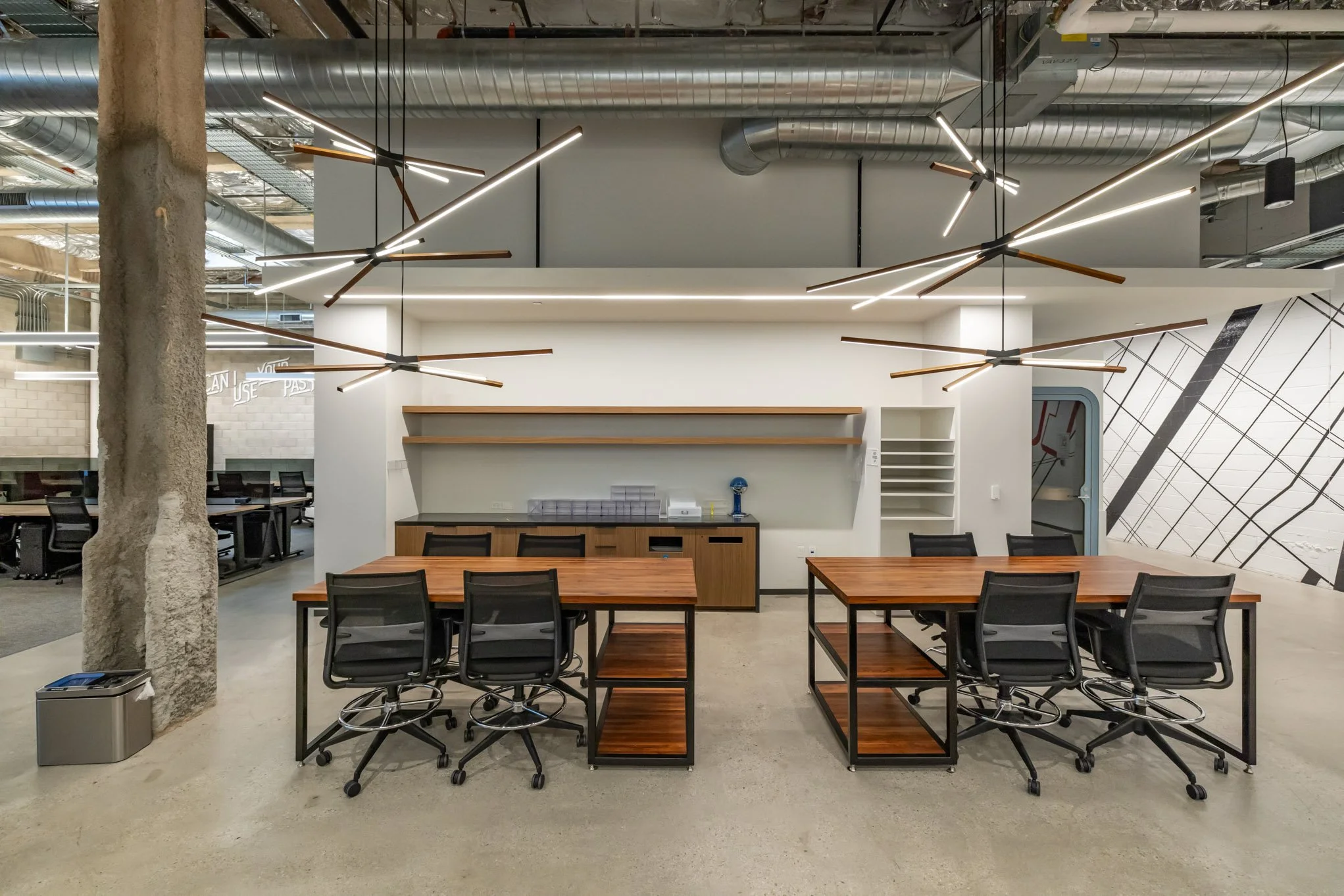 Modern office space with two wooden tables surrounded by black office chairs, a large wooden cabinet, open shelving, and unique ceiling lighting fixtures.