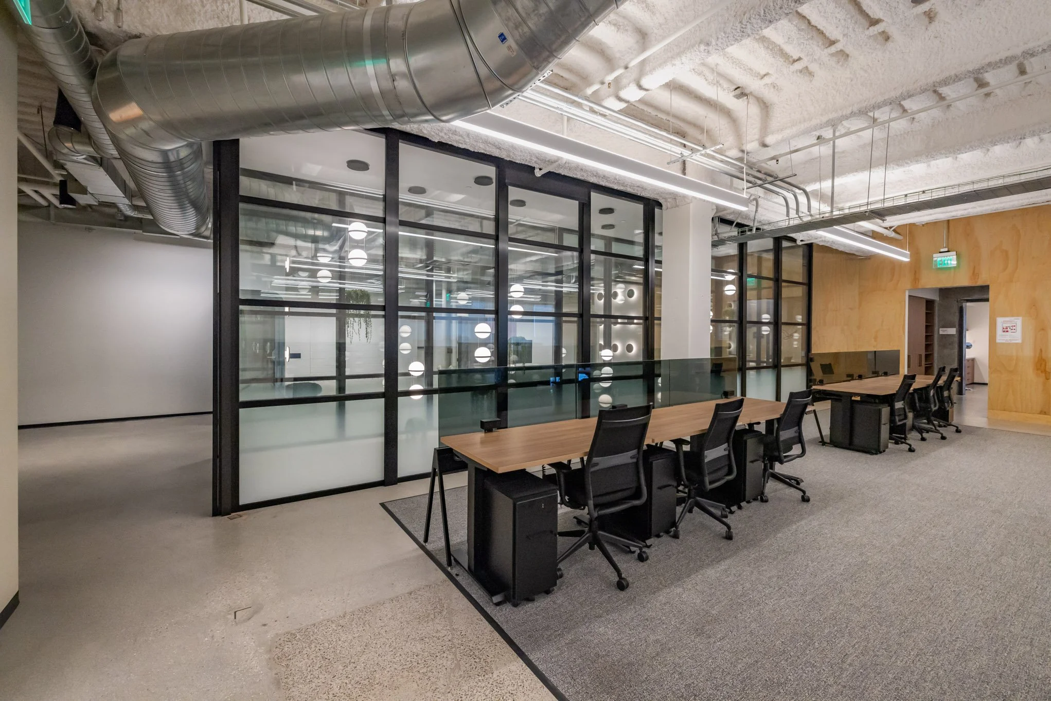 Office meeting area with black chairs, wooden tables, glass partitions, and visible industrial ceiling ductwork.