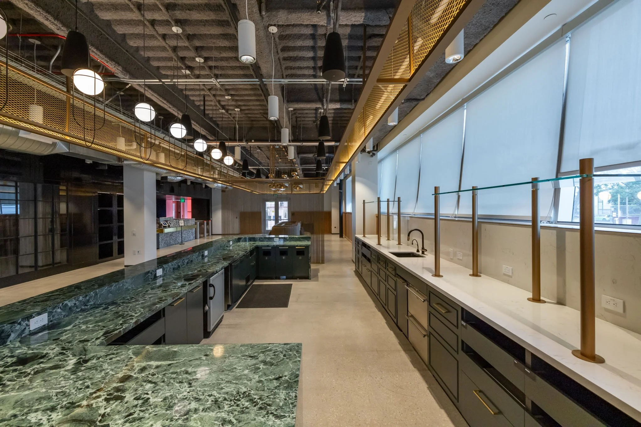 Empty modern kitchen with green marble countertops and black cabinets, large windows with shades, and industrial ceiling lighting.