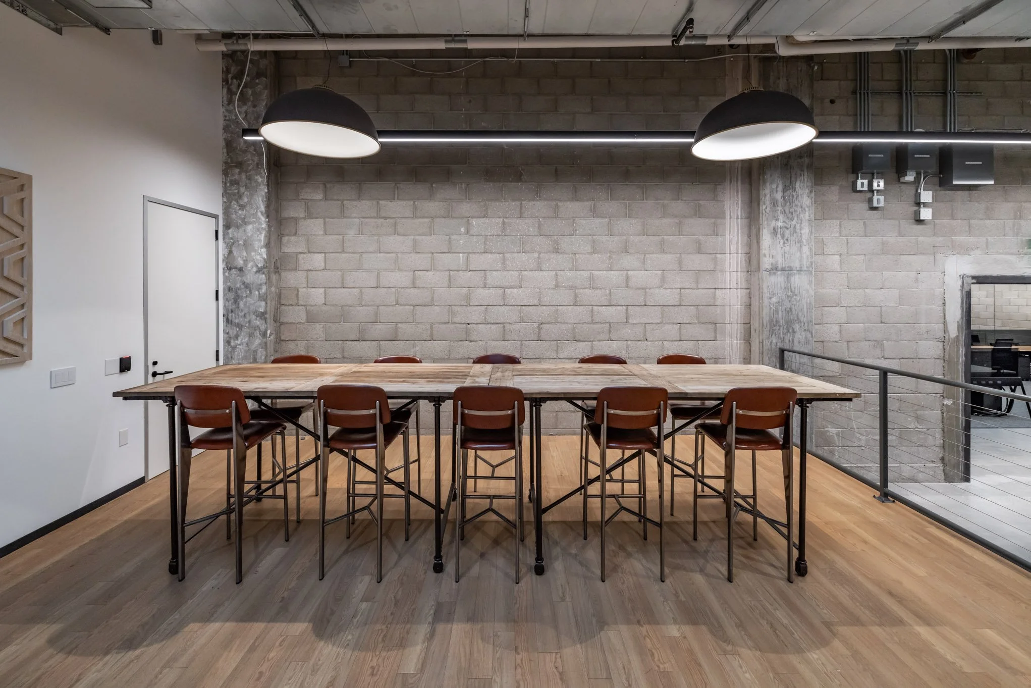 Meeting room with a long wooden table surrounded by ten brown chairs, exposed brick wall, modern black pendant lights, and wooden flooring.